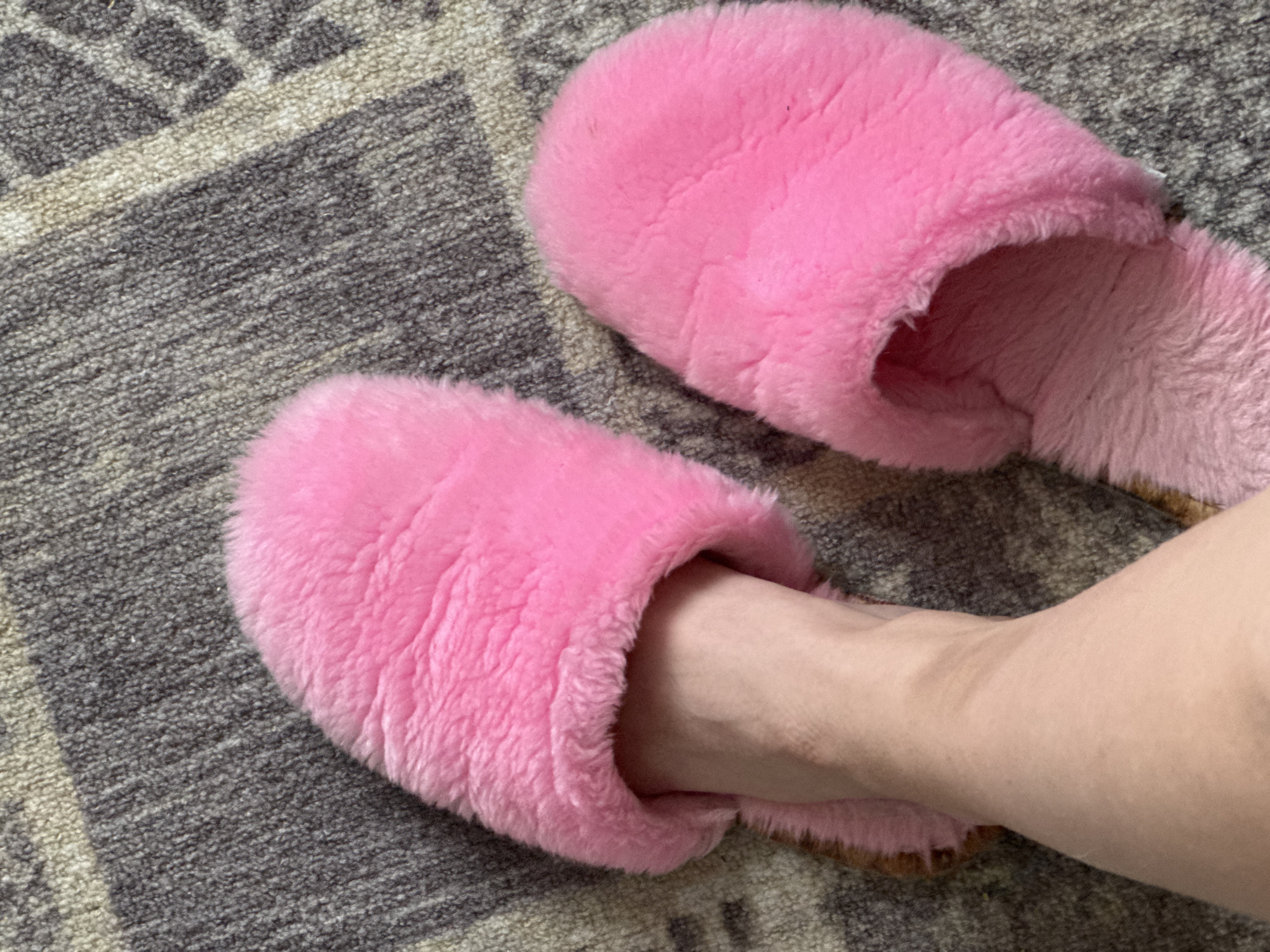 The image displays a close-up, high-angle view of a person's foot and two bright pink fluffy slippers resting on a patterned rug. One bare foot, with light-toned skin, is partially inserted into one of the soft, plush slippers, suggesting a moment of relaxation or the act of putting on/taking off footwear. The other identical pink slipper lies unoccupied beside it.
The slippers are the focal point, notable for their vibrant pink color and thick, shaggy texture, which conveys a sense of warmth and comfort. The soles of the slippers appear to be a light brown or tan shade.
The background is a textured rug or carpet patterned with shades of dark grey, grey, and light beige or off-white. The pattern is abstract or geometric, composed of intersecting lines and shapes. The setting is clearly indoors, indicative of a home environment in Izhevsk, Russia. No specific time of day, weather, or explicit emotions are discernible, but the scene generally evokes a feeling of domestic comfort. There is no visible text in the media file.