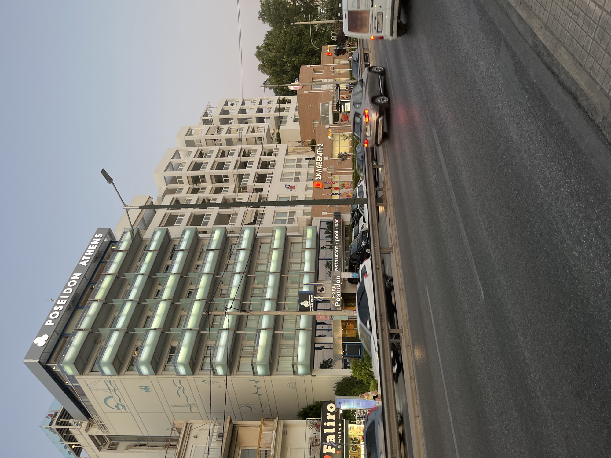The image captures a street scene in Paleo Faliro, Greece, dominated by the Poseidon Athens hotel and adjacent buildings. The Poseidon Athens hotel is a multi-story building with a modern design, featuring illuminated balconies and prominent signage. To its right, a white, multi-story building with a modern architectural style, characterized by numerous balconies and recessed windows, stands tall. In the foreground, a road with visible lane markings is occupied by several cars, some in motion, suggesting active traffic. A sidewalk with tiled paving is visible along the right edge of the road. Signs for "Faliro" and "Sklavenitis" are visible on the storefronts, indicating commercial establishments. The sky is a clear, pale blue, suggesting it is either early morning or late afternoon. The overall scene depicts a typical urban environment with a mix of commercial and residential buildings along a busy street.