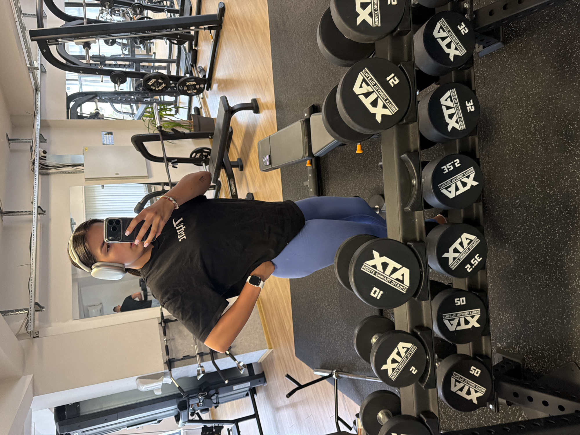 In a gym setting in Gabrovo, Bulgaria, a young woman with dark hair, wearing headphones, a black t-shirt, and blue leggings, stands and takes a selfie in a mirror. She is holding a smartphone in her right hand and has her left hand on her hip. Adjacent to her, a rack of dumbbells is visible, with weights ranging from 2 kg to 50 kg. In the background, various gym equipment, including weight machines and benches, are present. The flooring appears to be a combination of rubber mats and wooden planks.