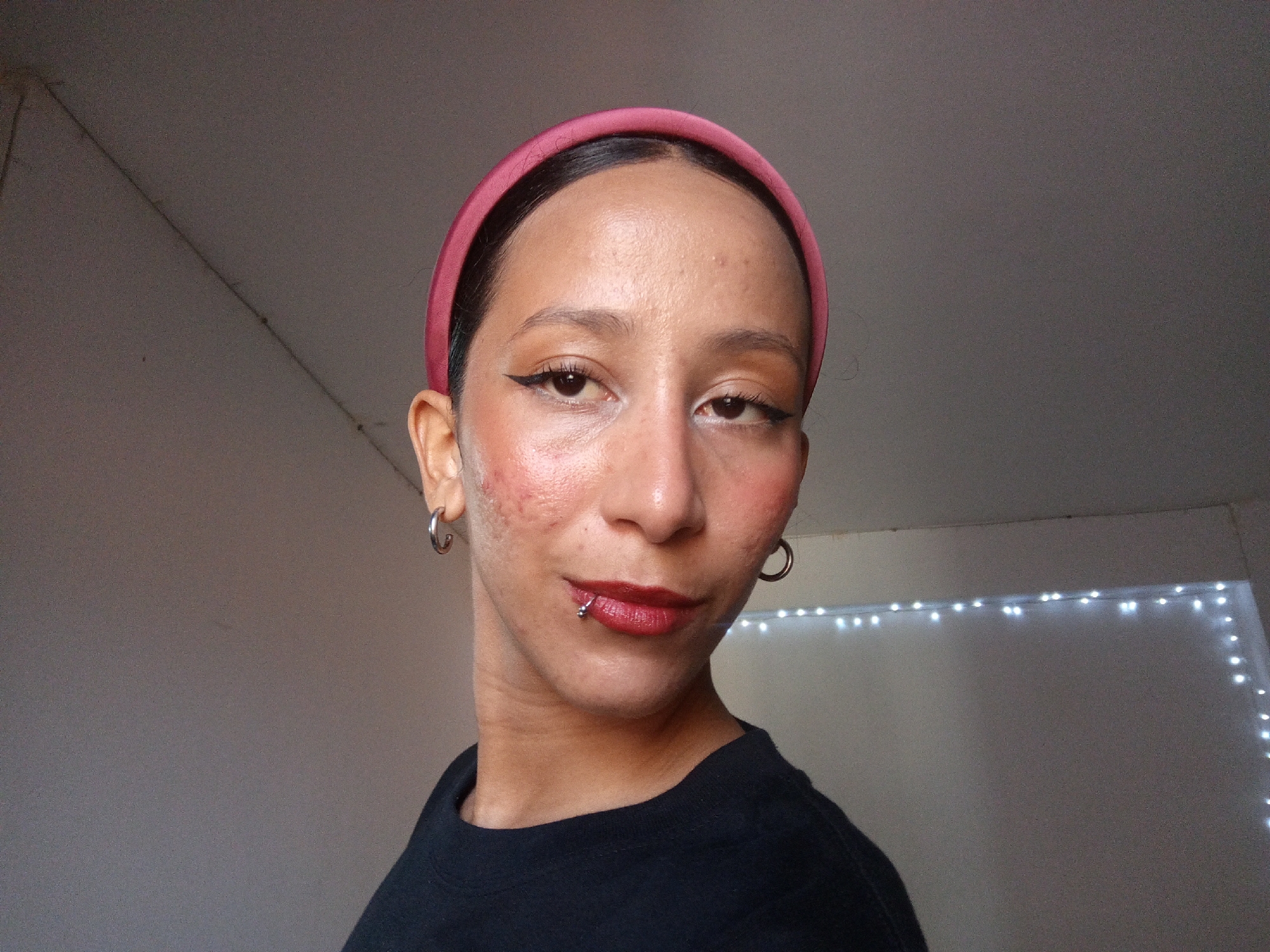 A woman with fair skin, wearing a dark shirt and a pink headband, poses indoors. She has dark hair pulled back and is wearing makeup, including eyeliner, blush, and lipstick. She has a lip piercing on the left side of her lower lip and is wearing hoop earrings. The background consists of a light gray wall and ceiling, with a string of lights visible in the lower right corner. No specific information confirms the location in the city of Porlamar, Venezuela.