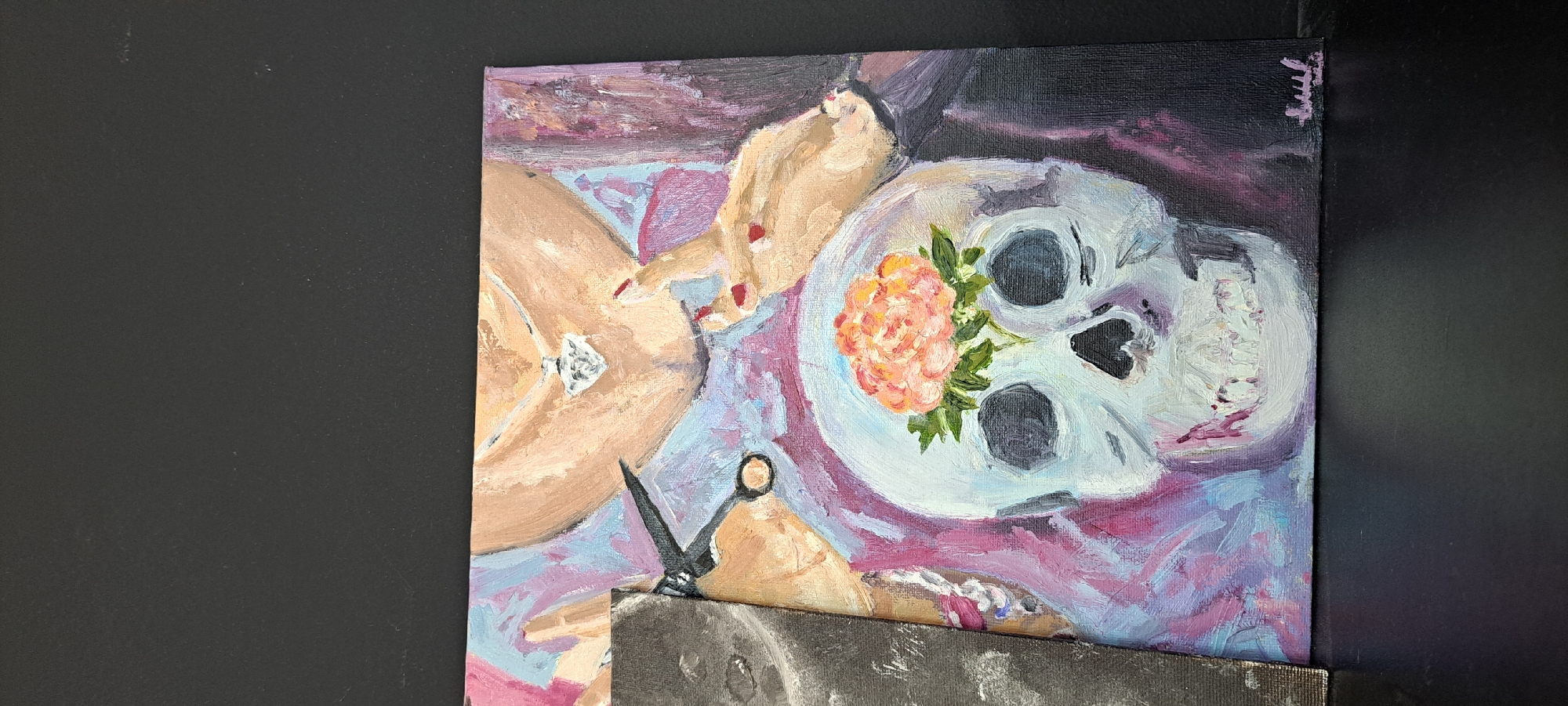 The image shows a close-up of a painting. The painting depicts a white skull with a pink and orange flower on top of its head. A pair of scissors is held over the skull. A person's hands, with red nail polish, are visible in the upper right, and a diamond necklace is worn around the neck. The painting is rendered in an impressionistic style with visible brushstrokes. The background is dark. The painting is positioned on a dark surface. The bottom portion of the image shows a black surface, possibly a table or shelf, with a dark, out-of-focus background above it. There is no indication of the city of Yessentuki, Russia, within the image.