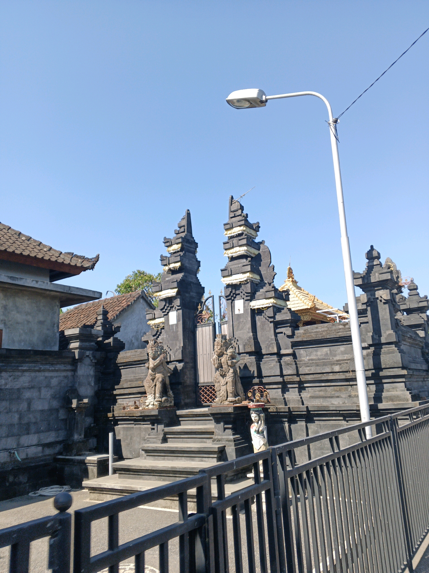 The image depicts a temple entrance with traditional Balinese architecture under a clear blue sky. The temple features dark grey stone structures with tiered roofs, adorned with golden accents. Statues of guardians flank the entrance, and a wide set of stone stairs leads up to the gate. A modern lamppost stands to the right of the temple, along with a black metal fence in the foreground. To the left, a building with a tiled roof and grey walls is partially visible. The scene suggests the temple is located in Abiansemal, Indonesia, a region known for its rich cultural heritage and numerous temples.