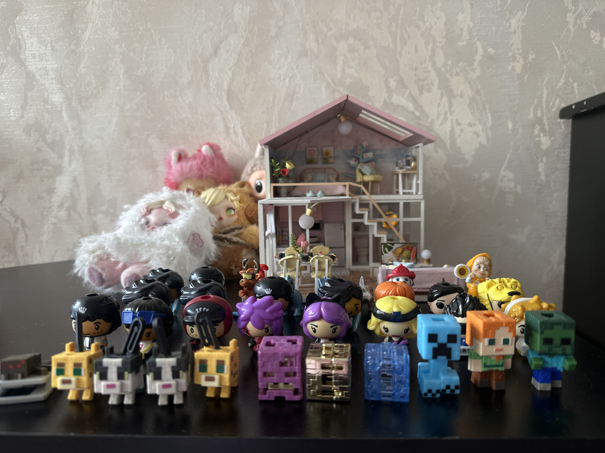 The image shows a collection of toys arranged on a dark, reflective surface, likely a table. In the foreground, there is a row of small Minecraft figures of various characters and blocks. Behind the Minecraft figures are slightly larger figures with stylized faces and helmets. Behind these figures, a dollhouse with a pink roof and detailed interior is visible. To the left of the dollhouse are several plush toys. The background is a light-colored wall with a textured pattern. To the right, a black piece of furniture is partially visible. The image does not contain specific details to definitively confirm its location as Yerevan, Armenia.