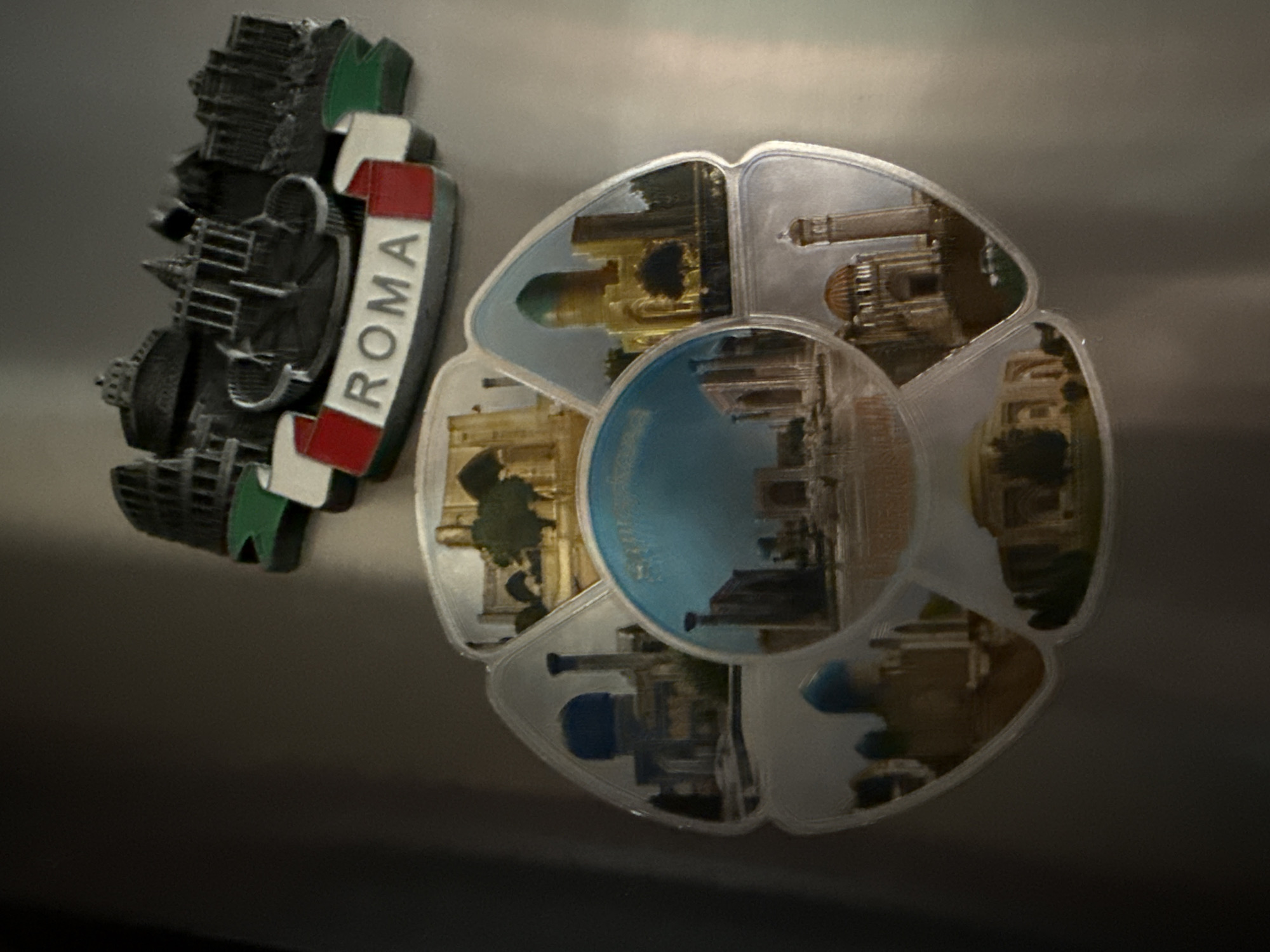 This media file showcases two refrigerator magnets. The magnet on the left is a black, three-dimensional replica of the Colosseum in Rome, with the word "ROMA" inscribed in white letters on a red and white banner. It also features elements reminiscent of the Italian flag.
The magnet on the right is a novelty item shaped like a flower or a pinwheel, with each petal displaying a different image. The central part of this magnet features a circular image with a blue sky and the reflection of buildings. The surrounding petals show various architectural landmarks, possibly in Tashkent, Uzbekistan, given the provided location context. These images include golden-domed buildings, intricate facades, and trees, suggesting an outdoor setting. One petal clearly shows a blue dome, and another depicts a structure with multiple arches.
The overall scene is a close-up of these two magnets attached to a reflective, metallic surface, likely a refrigerator door. The lighting is somewhat dim, casting shadows and creating a slightly moody atmosphere. There are no people visible in the frame. The focus is entirely on the magnets themselves and their intricate details. The visible text on the left magnet is "ROMA".