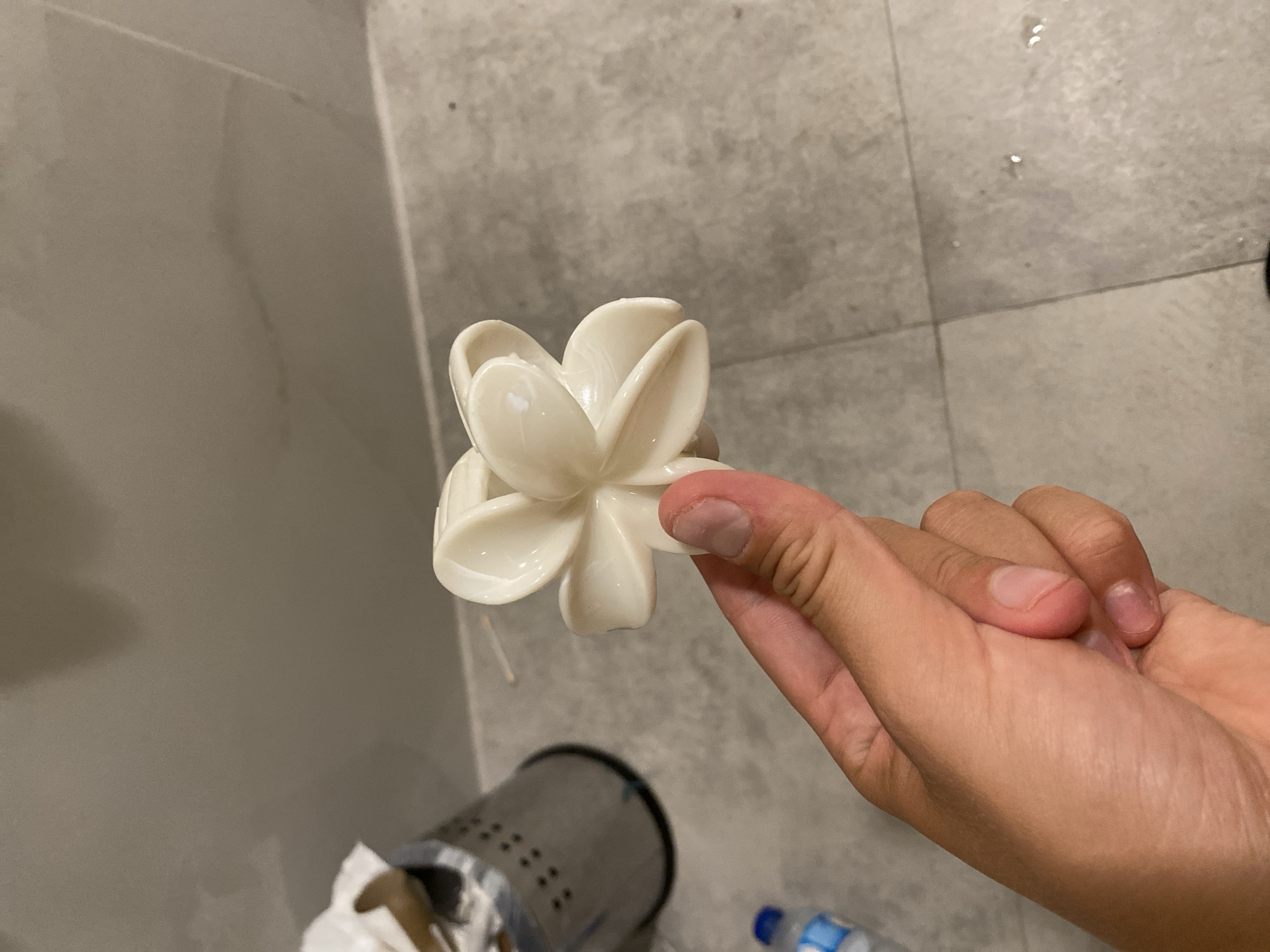 A person's hand is holding a white, flower-shaped object against a tiled floor and wall. In the background, a metal trash can with holes is visible. A partially filled plastic water bottle is also present. The lighting suggests an indoor environment. The location in Aksu, Turkey is not discernible from the image.