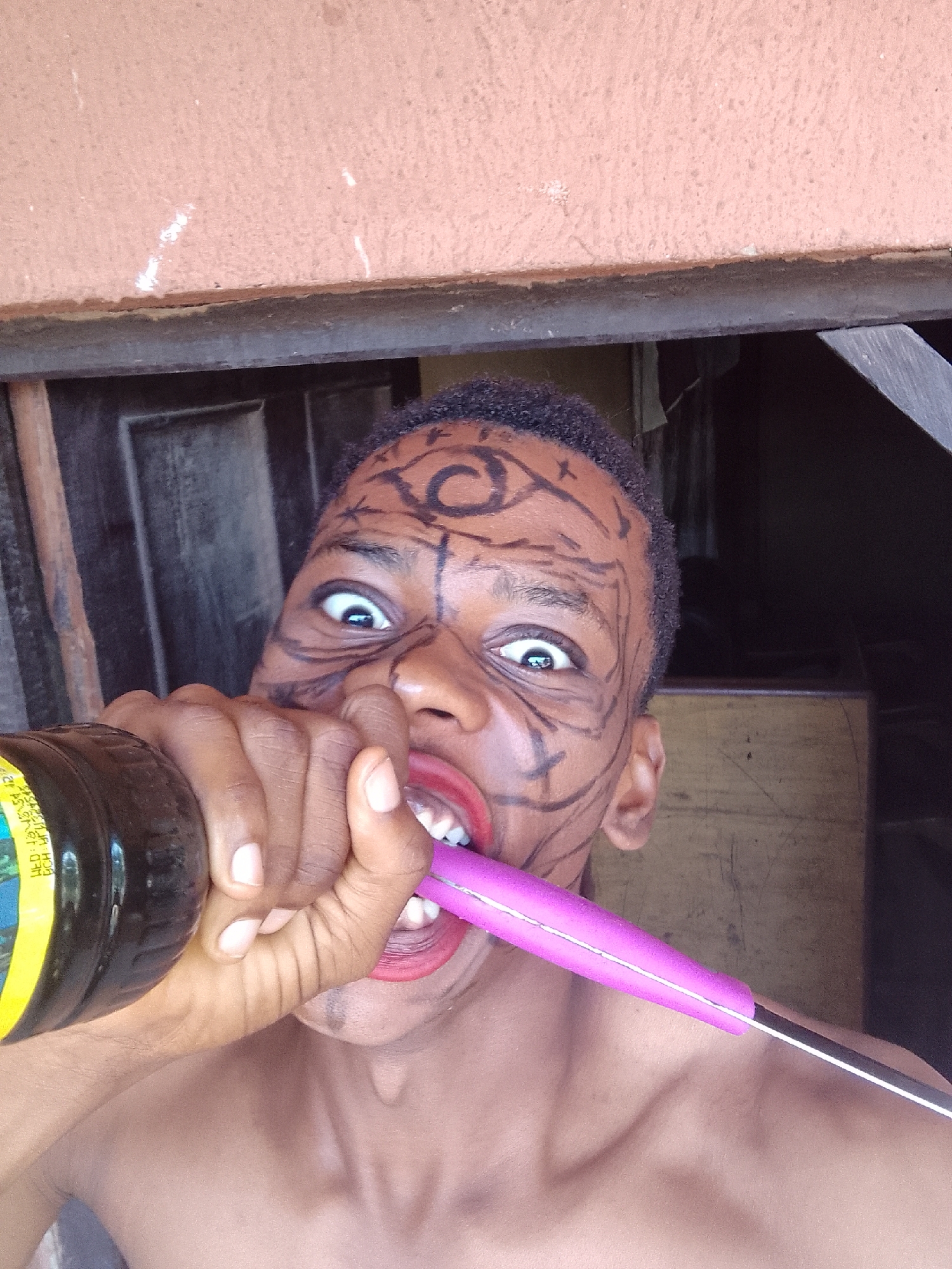 A shirtless person with brown skin and a painted face is holding a dark bottle to their mouth with their left hand and a pink-handled knife in their teeth. The person's face has dark, painted designs, including swirls on their forehead and lines on their cheeks and under their eyes. The person's eyes are wide. The background is out of focus, but appears to be an interior space with a dark doorframe above the person. The location is Unknown, Unknown.