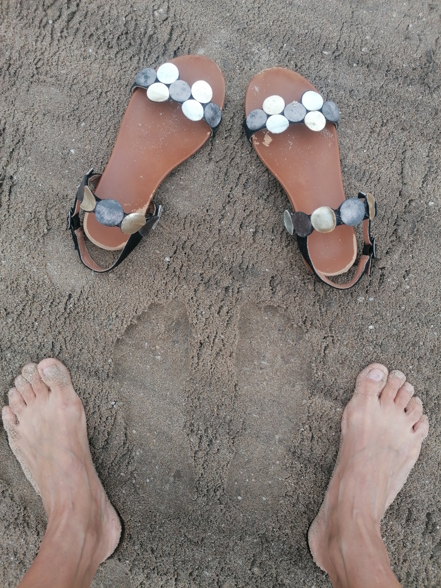An overhead shot captures a pair of bare feet and a pair of decorative sandals on light brown-grey sand. The sandals are positioned at the top of the frame, with the bare feet placed below them, slightly spread apart.
The sandals feature brown soles and black straps. Across the front of each sandal, a decorative band is adorned with several flat, circular embellishments in varying metallic shades of silver, grey, and bronze/gold. The left sandal and left foot are on the left side of the image, and the right sandal and right foot are on the right. Both feet are lightly covered in sand, particularly around the toes.
The sand exhibits a fine, granular texture. Between the sandals and the feet, faint impressions in the sand are visible, suggesting previous footprints. The scene implies a moment of leisure, perhaps at a beach or sandy area where the individual has just removed their sandals or is about to put them on. This activity takes place in a sandy environment, consistent with a coastal location like Bai Chay Ward, Vietnam. No text is visible in the image.