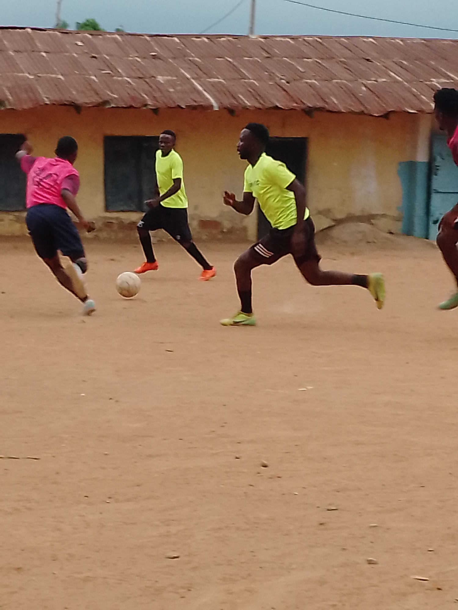The image shows a soccer game in Akwanga, Nigeria. Four men are visible on a dirt field. One man in a pink jersey is kicking a soccer ball, while another man in a bright yellow jersey runs to intercept. Another player in a yellow jersey is standing back, awaiting the play. The partial image of a fourth player is visible at the right edge of the frame, also wearing pink. Behind the players is a low building with a corrugated metal roof and two square windows. The sky is overcast.