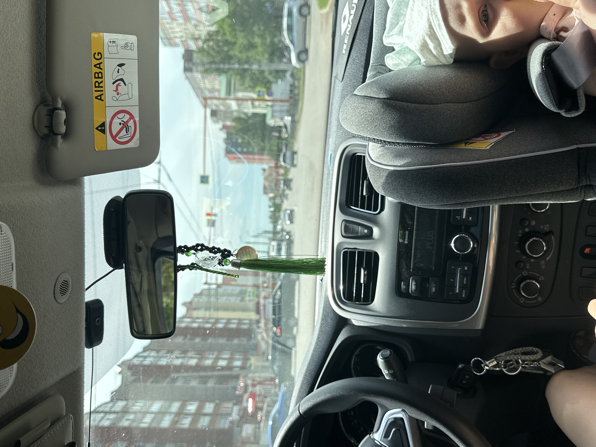 The image captures a view from the passenger seat of a car driving through the city of Sterlitamak, Russia, on a cloudy day. The dashboard is visible, featuring the steering wheel, air vents, radio, and climate control knobs. A car seat with a baby is positioned to the right, with the baby's face partially visible. A keychain with several keys hangs from the ignition. Outside the car, apartment buildings and trees line the street, with other vehicles and traffic signals indicating a typical urban scene. A sun visor with an "AIRBAG" warning sticker is also visible on the upper right.