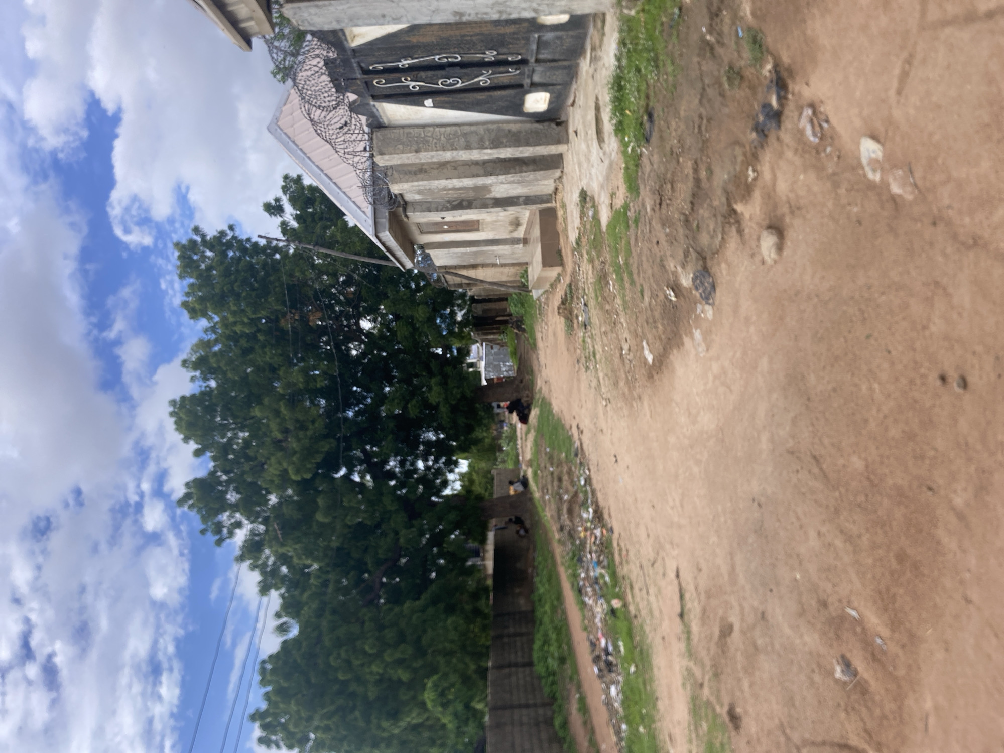 The image depicts an unpaved street or path in a residential or semi-urban area within the city of Unknown, Unknown, under a partly cloudy sky. The upper portion of the frame shows a blue sky with scattered white cumulus clouds; multiple power lines extend horizontally across the upper-left quadrant.
On the left side, a long, dark grey concrete block wall runs parallel to a dirt path. Adjacent to this path, a significant accumulation of litter, consisting of plastic bags, bottles, and general debris, is present on the ground. Further back, beneath the canopy of large green trees, several indistinct human figures are seated or resting on the ground.
On the right side, a building with a light-colored, possibly corrugated, pitched roof is visible. The roof is topped with coils of razor wire, indicating security measures. The building's facade is constructed of concrete, featuring prominent vertical decorative or structural elements and a single small, dark wooden window. In the foreground, a large, dark metal gate with ornate scrollwork and geometric patterns stands open, connected to the building's perimeter.
The ground surface throughout the foreground and mid-ground is predominantly reddish-brown dirt, appearing uneven and marked by small rocks, sparse patches of green grass, and scattered additional litter. Another indistinct human figure is seated under a tree in the mid-ground on the far right.