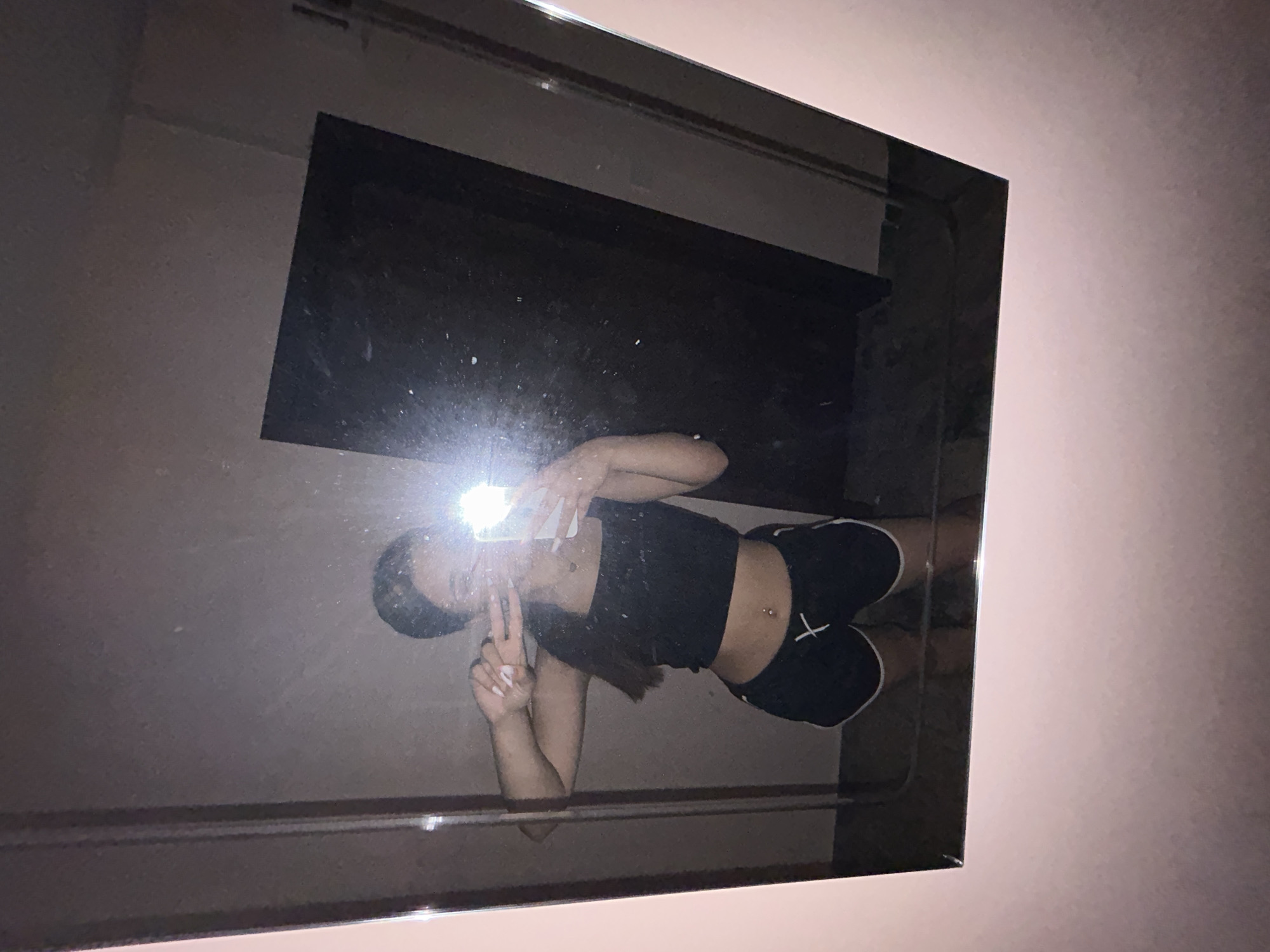 In a dimly lit room, a person is taking a mirror selfie. They are wearing a black crop top and black shorts with white trim. The person is holding up a smartphone in their right hand, taking the picture, and is making a peace sign with their left hand. A bright flash from the phone illuminates the scene. The background appears to be a plain wall with a dark, rectangular object, possibly a door or a closet, to the right. The mirror has a pale pink frame. The image does not provide enough information to determine the location in the city of Shumen, Bulgaria.