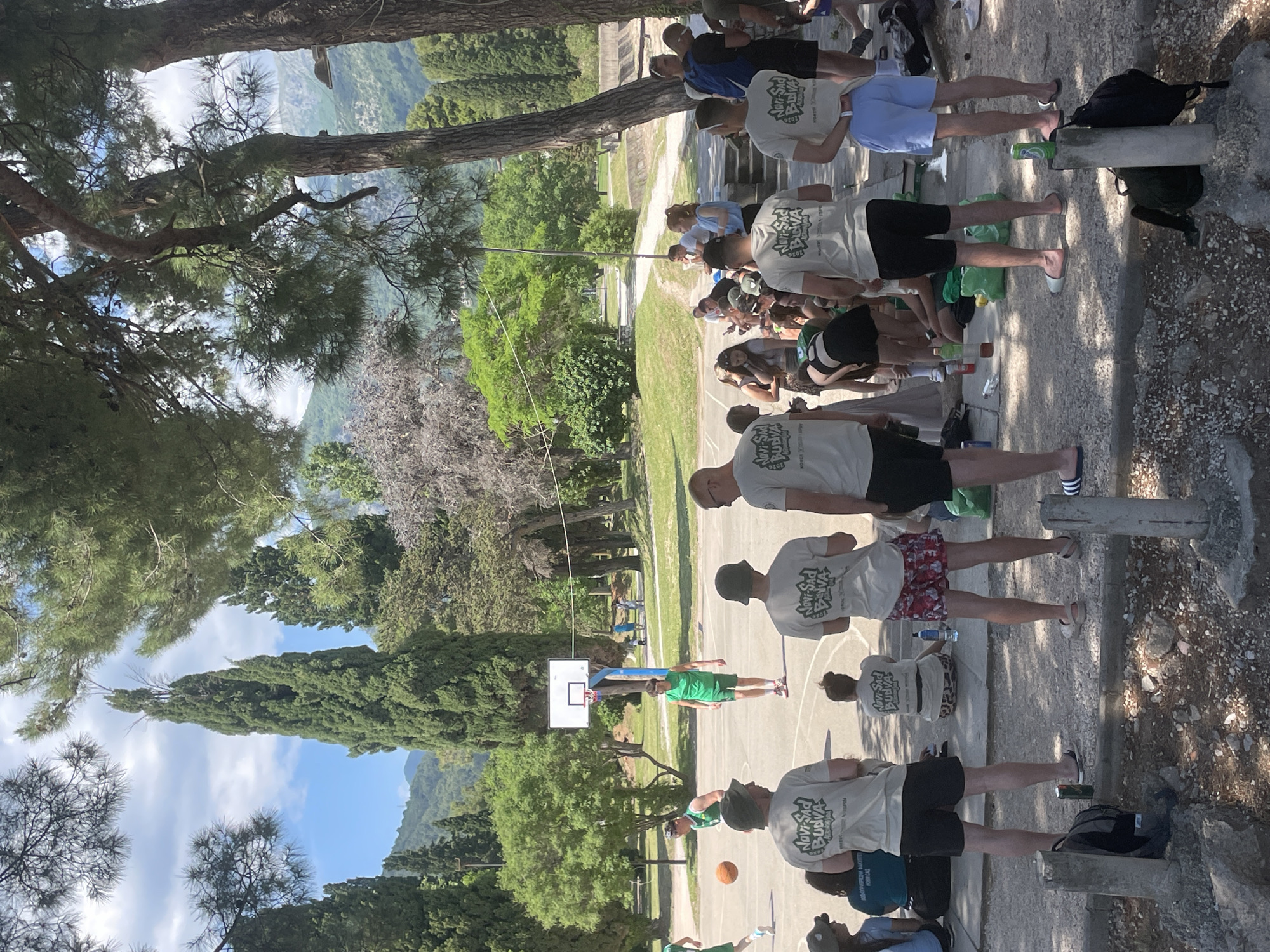The image shows a group of people, presumably associated with an event or gathering ("Novi Sad Budva" is printed on their shirts), outdoors in what appears to be a park-like setting. The group includes at least ten people who are lined up. Some are standing while others are seated. There's a basketball court visible in the background, suggesting the location is near recreational facilities. The environment includes trees, greenery, and possibly mountains or hills in the distance.