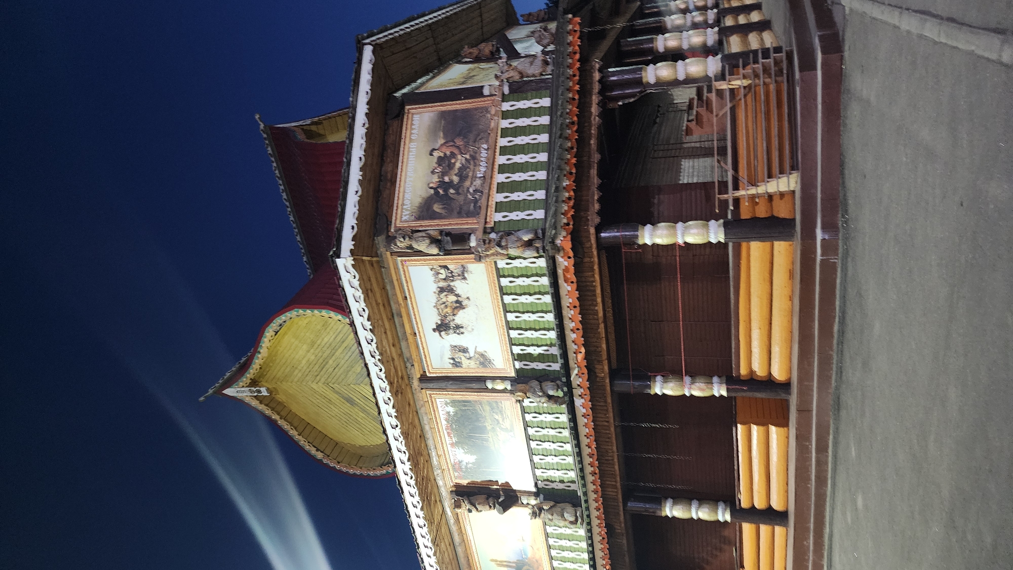 The image shows a traditional Russian wooden building, possibly a pavilion or a small museum, located in Moscow, Russia. The building features ornate wooden carvings, colorful paintings displayed on its facade, and a distinctive onion-shaped roof. The structure is illuminated, suggesting it is evening or night. The surrounding environment is dark, with a deep blue sky visible above, and there is a slight light streak across the sky, possibly from a distant light source or a vehicle. The lower part of the building has wooden railings and what appear to be closed shutters or doors, indicating it might be closed to the public at the time the photo was taken. A staircase leads up to the main level. The overall impression is of a well-maintained, culturally significant architectural piece.