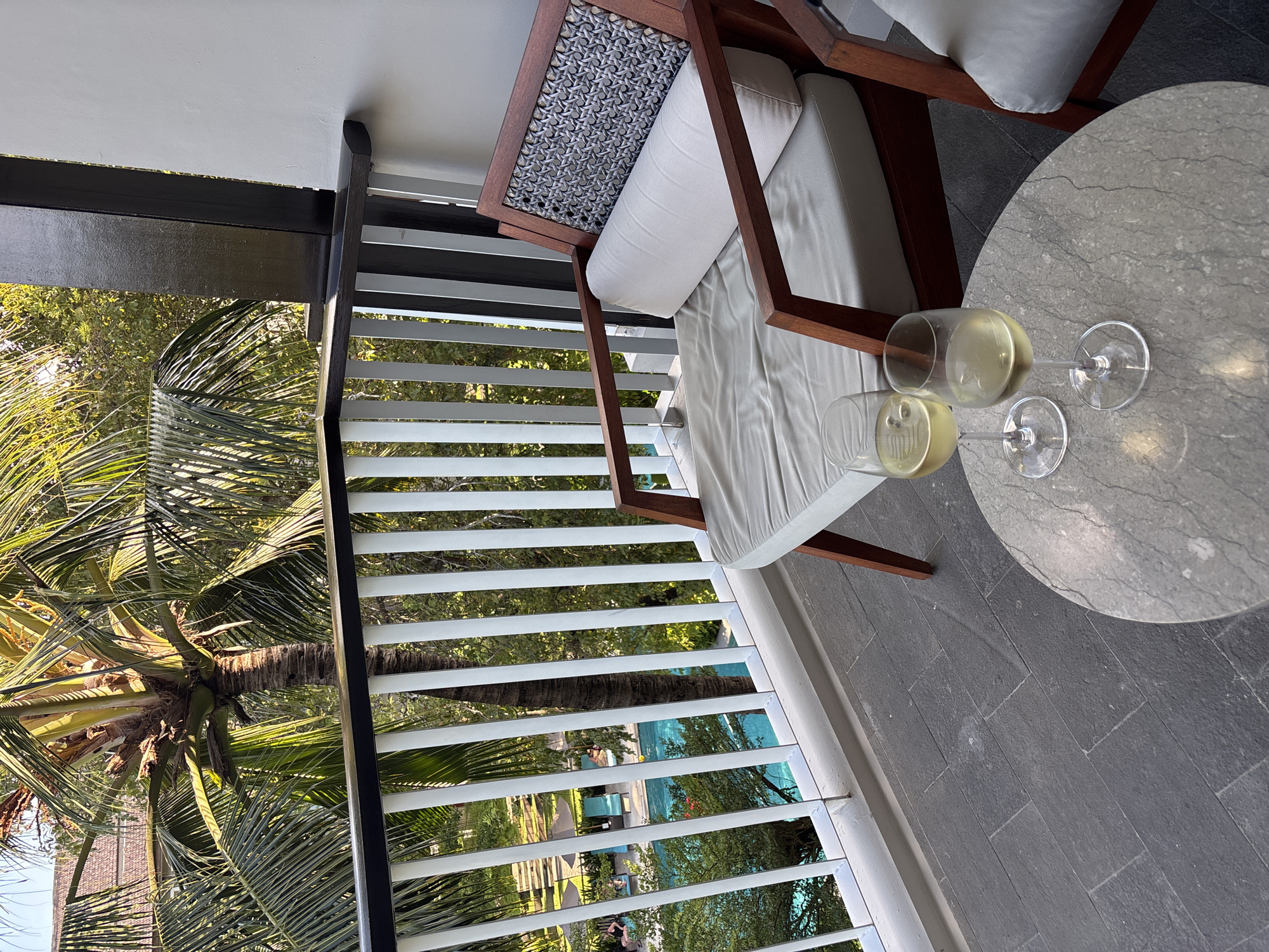 This image captures a serene outdoor scene from a balcony or patio, likely at a tropical resort in Kuta Selatan, Indonesia. In the immediate foreground, a round, grey marble-patterned table holds two stemmed glasses, each containing a light yellow beverage, possibly white wine or a cocktail. Adjacent to the table, an inviting outdoor armchair features a dark wooden frame, plush white cushions, and a distinctive grey woven pattern on its backrest.
A white horizontal slatted railing separates the seating area from the lush view beyond. Through the slats, a vibrant tropical landscape is visible, dominated by numerous large palm trees and other dense green foliage. Dappled sunlight filters through the leaves, indicating a bright and clear day, possibly late afternoon or early morning. In the distance, a glimmering blue swimming pool can be seen, surrounded by pathways and more greenery. A solitary figure, dressed in dark clothing, is partially visible near the pool area. The balcony floor is made of dark grey rectangular tiles. The overall scene evokes a sense of relaxation and a luxurious vacation setting. No discernible text is visible in the image.