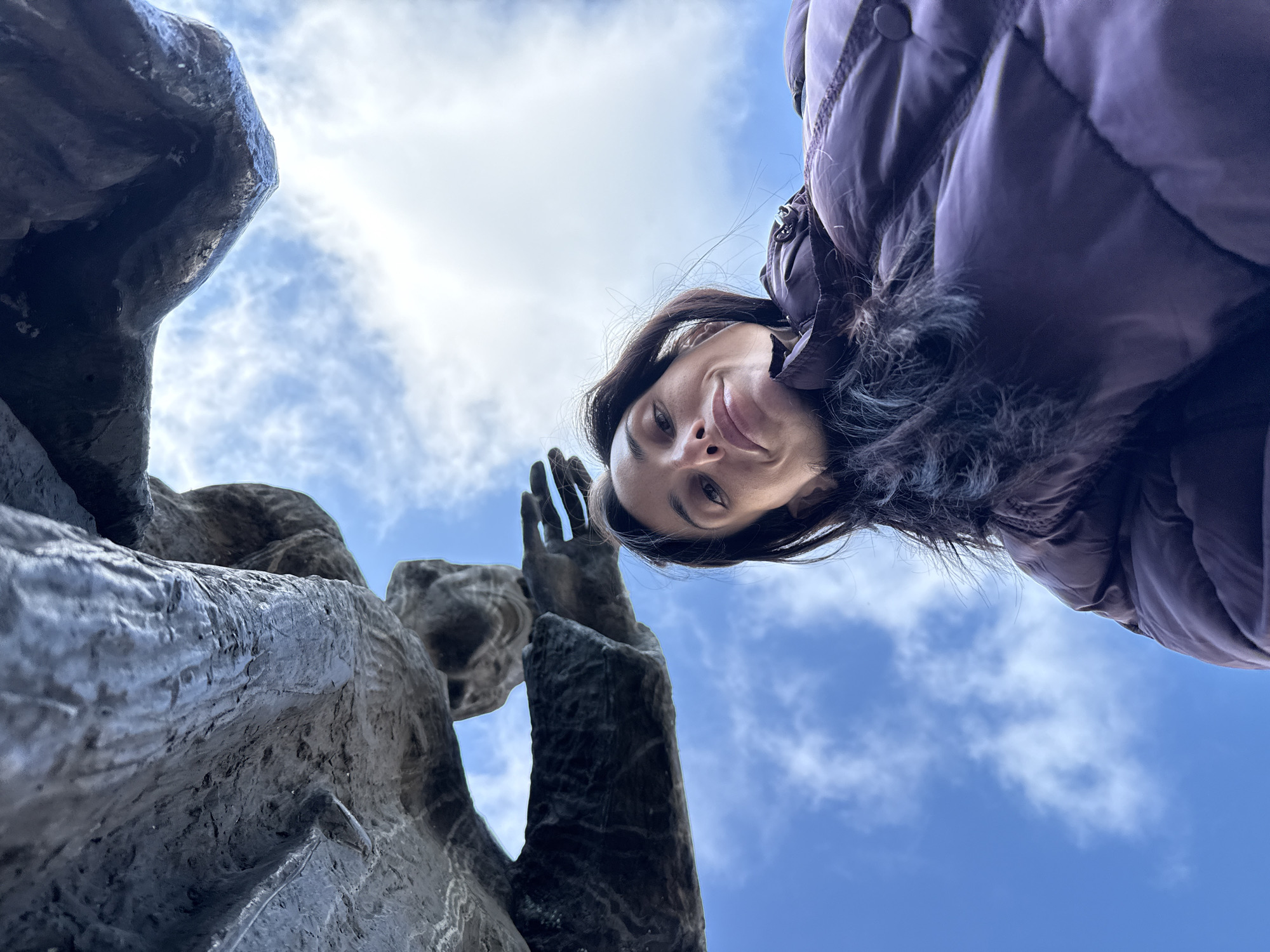 A woman takes a low-angle selfie with the monument "Broken Chains" in Pleven, Bulgaria. She is wearing a dark purple winter coat. The monument is made of dark stone and depicts a figure with raised hands. The sky is blue with scattered white clouds.