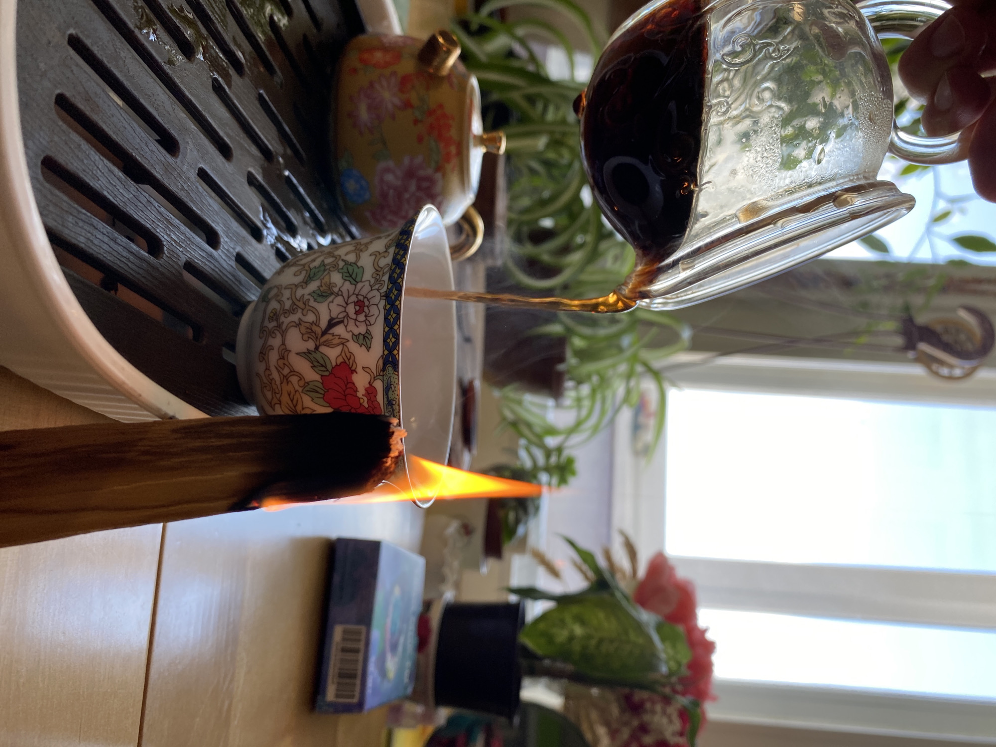 A brightly lit, rotated indoor shot shows tea being poured from a clear glass pitcher into a floral teacup on a dark brown wooden slatted tray with a white rim. To the left of the teacup is a burning stick with a visible flame. A colorful teapot sits on the tray behind the teacup. Behind the tray and tea arrangement, there is a variety of houseplants in front of a window. A small blue box and other decorative items sit on the surface below the tray. The image lacks any evidence to suggest the location of the image as Chaikovsky, Russia.