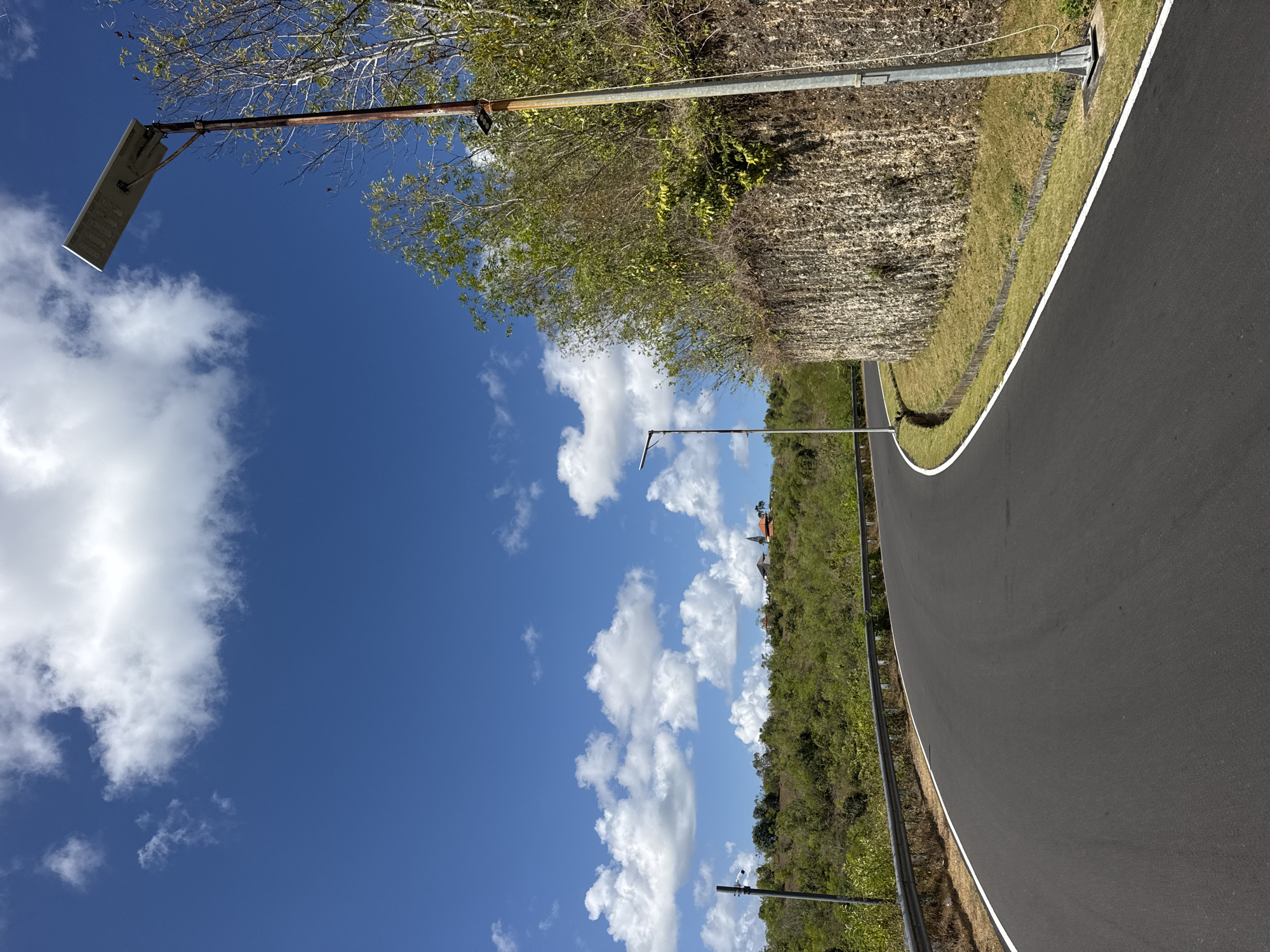 The image shows a paved road curving to the left through a green, hilly landscape. The road has white lines marking its edges. On the right side of the road, a rough-hewn stone wall rises, topped with dense green foliage. A tall, rusty lamppost stands near the wall, its solar panel tilted towards the sky. In the distance, on top of a hill, a small cluster of buildings, one with a red roof and a steeple, is visible. The sky is a vibrant blue, dotted with fluffy white clouds. The scene appears to be located in Kuta Selatan, Indonesia, judging by the lush vegetation and architectural style of the distant buildings. The overall impression is of a scenic, semi-rural environment under a bright, sunny sky.
