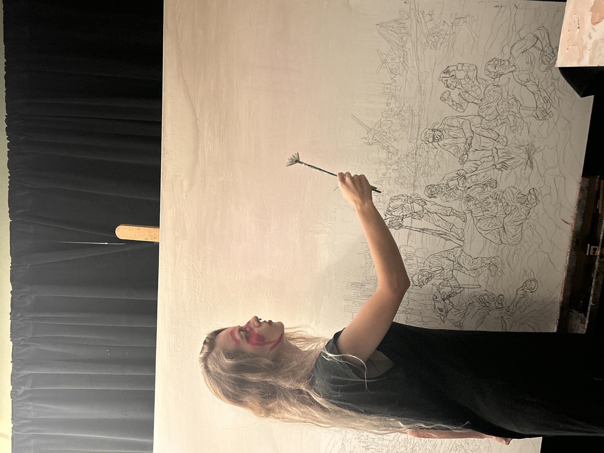 The image shows a fair-skinned person with long blond hair, wearing a black shirt and red markings on their face, standing in front of a large white canvas. The person is holding a paint brush towards the upper portion of the canvas. The canvas has a detailed sketch of figures engaged in what appears to be a battle scene. The canvas is on an easel and there is a palette with paint visible to the right. To the left of the canvas is a black curtain. The location is not identifiable as Cape Coral, United States of America from the given visual information.