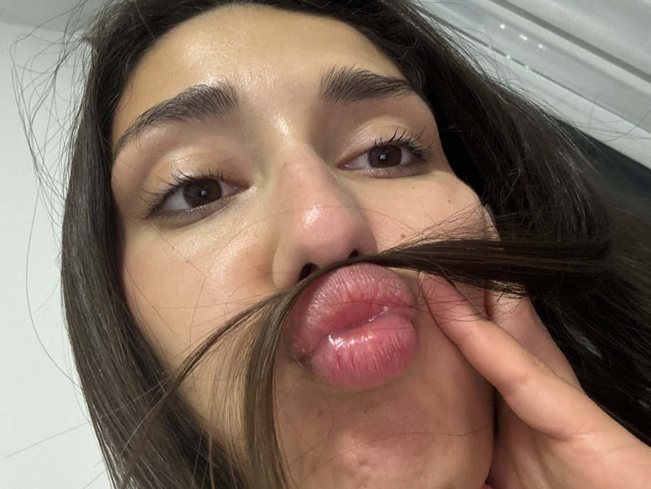 A woman holds hair under her nose, mimicking a mustache. She looks up at the camera with a playful expression. Her lips are slightly puckered, revealing a glossy finish. Her skin has a dewy texture and her eyebrows are well-groomed. The background is a blurred light color.