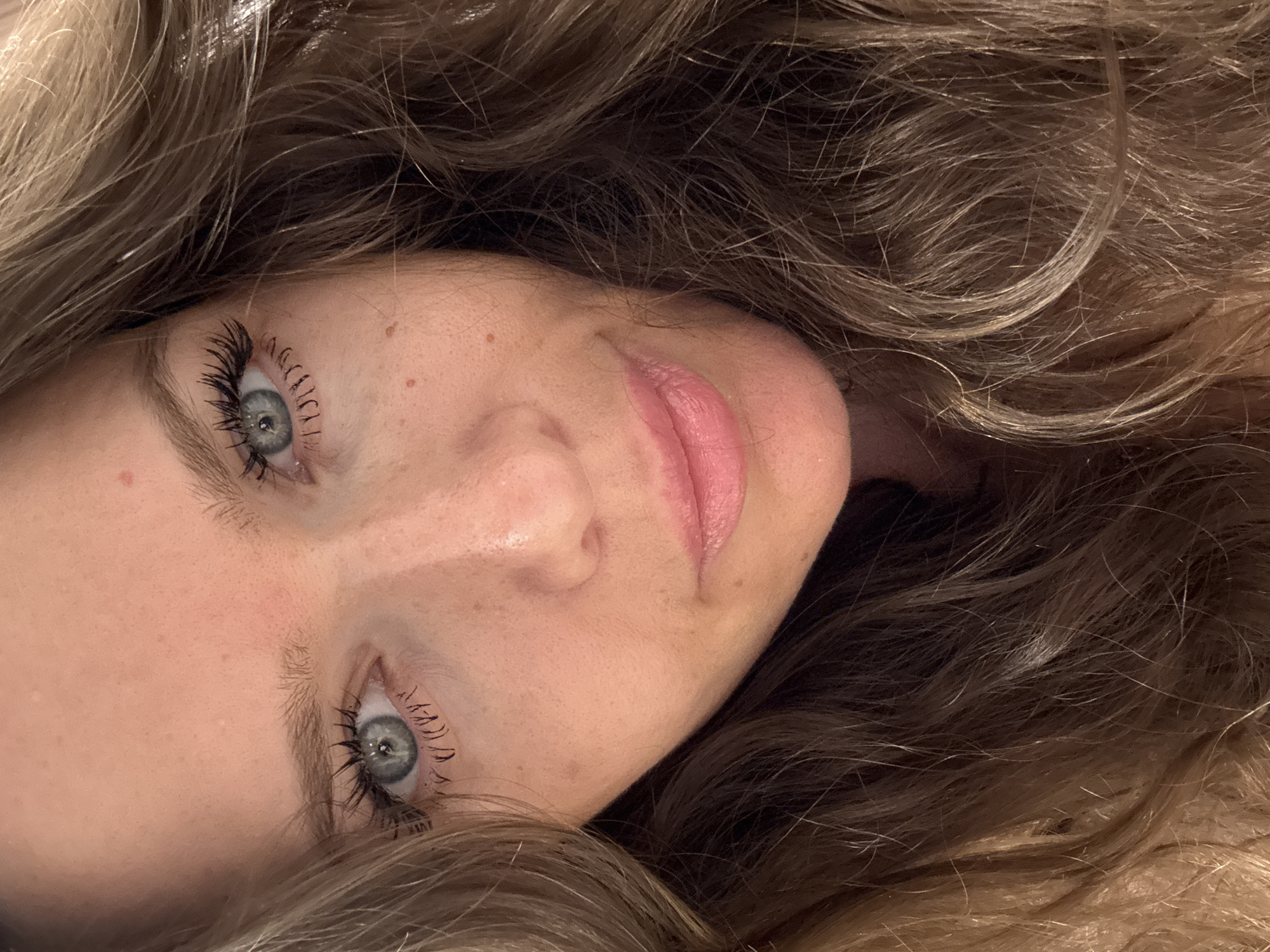 This media file is a close-up, vertical, personal portrait of a young Caucasian woman's face, with a shallow depth of field. The woman has fair skin with a smattering of freckles, light blonde wavy hair, and light blue eyes. Her lashes are dark and appear to be coated with mascara. She is wearing light pink lipstick, and her lips are slightly parted. Her expression is neutral, and the lighting suggests it may be indoors, possibly in a dimly lit room. The image is a self-portrait, as indicated by the perspective. There is no visible text in the image. The scene appears to be intimate and personal, with the focus solely on the woman's face and hair. There are no external environmental cues to suggest a specific location or time of day beyond general indoor lighting. The image is candid in nature, capturing a moment of quiet repose.