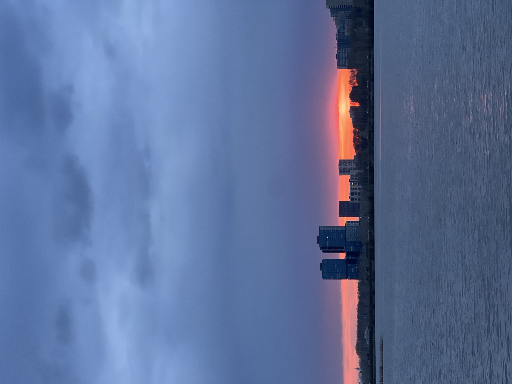 The image shows a sunset in Kazan, Russia. The sky is a mix of dark blue and orange, with the sun setting over a body of water. Buildings are visible along the shoreline. The water reflects the colors of the sky, creating a mirror effect. There are no people visible in the image.