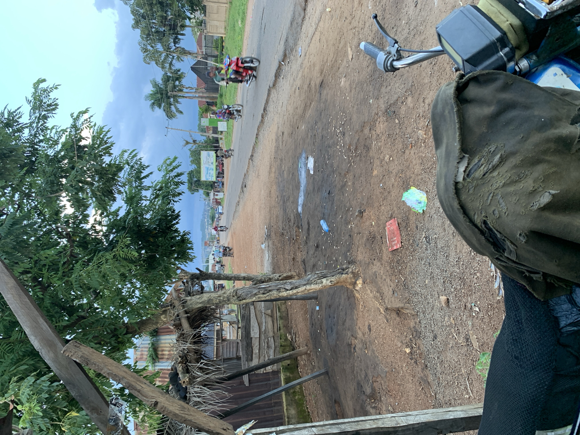 The image depicts a roadside scene in the city of Kabba, Nigeria. In the foreground, a portion of a damaged motorcycle is visible, including the handlebar, seat, and part of the fuel tank. The seat is covered with a tattered, olive-green fabric. The ground is unpaved, consisting of reddish-brown dirt and scattered debris, including a discarded plastic bottle and a red rectangular object.
To the left of the foreground, there is a makeshift structure with a thatched roof supported by wooden posts and corrugated metal walls. Behind this structure, a few trees with green foliage are present.
Further down the unpaved roadside, a paved road stretches into the distance. Several individuals on motorcycles are traveling along this road. Some buildings and palm trees are visible on the right side of the road. The sky is overcast with clouds. The scene appears to be in a rural or semi-urban area within Kabba.