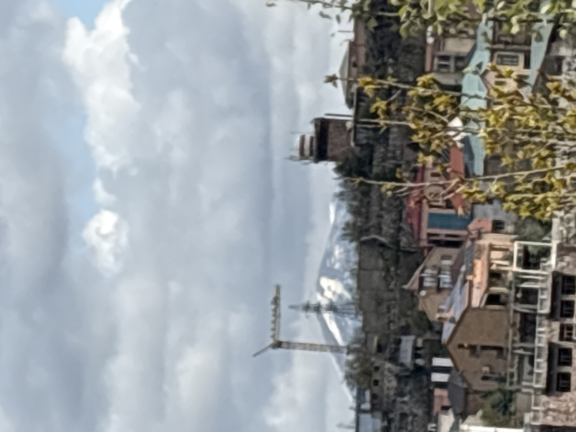 The image, correctly oriented, presents an expansive daytime view of an urban area in Yerevan, Armenia, set against a mountainous background. The sky, occupying the upper portion of the frame, is a mix of bright white and grey clouds, with patches of clear blue sky visible between them, indicating partly cloudy weather.
In the mid-ground and foreground, a dense collection of buildings forms the urban landscape. These structures vary in size, color, and architectural style, featuring different rooflines and facades, suggesting a blend of residential and commercial properties. Some buildings have multiple stories, and details like windows are faintly visible. Trees with green and yellowish foliage are scattered throughout the foreground, partially obscuring some of the structures and adding a natural element to the scene.
A notable object in the mid-ground is a tall, yellow construction crane, its boom extended, indicating ongoing development or building activity in the area.
Dominating the distant background is a majestic, snow-capped mountain range, its peaks appearing stark white against the cloudy sky. The presence of these mountains is a strong geographical indicator for the region.
The time of day appears to be during daylight hours, likely mid-day, given the diffused light and lack of strong shadows. No people are visible, and no specific activities are clearly discernible other than the implied construction. There is no visible text in the image. The scene conveys a sense of a city nestled within a dramatic natural environment, with continuous urban growth.