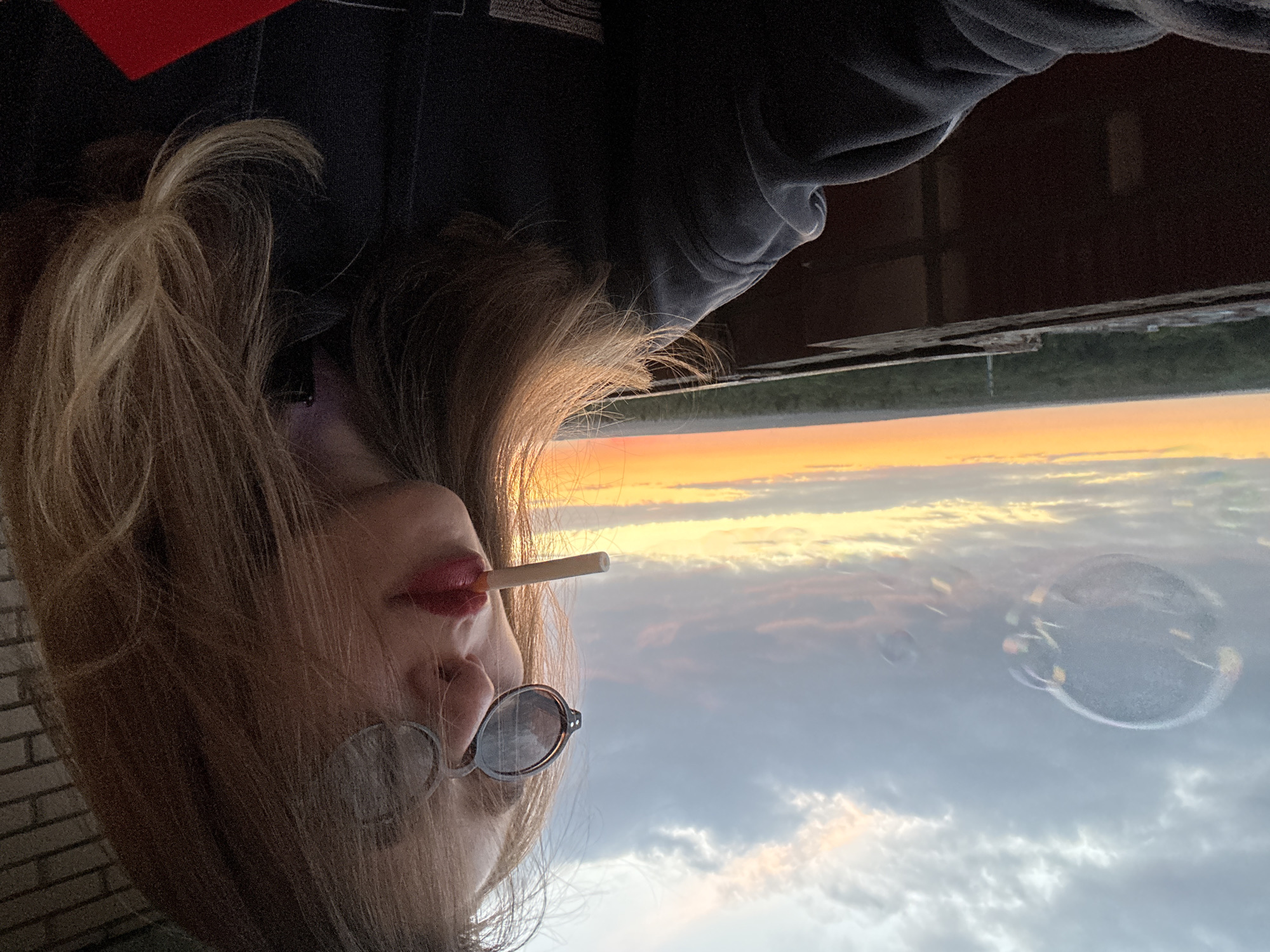 A medium shot captures a young woman with blonde hair and sunglasses holding a cigarette in her mouth, looking to the right. She is wearing a dark hoodie. The background consists of a cloudy sky with an orange and yellow sunset, and a distant view of trees and buildings. A large, translucent bubble floats in the sky to the left of the woman. The scene appears to be an outdoor setting, possibly a rooftop or balcony, given the railing and distant cityscape.