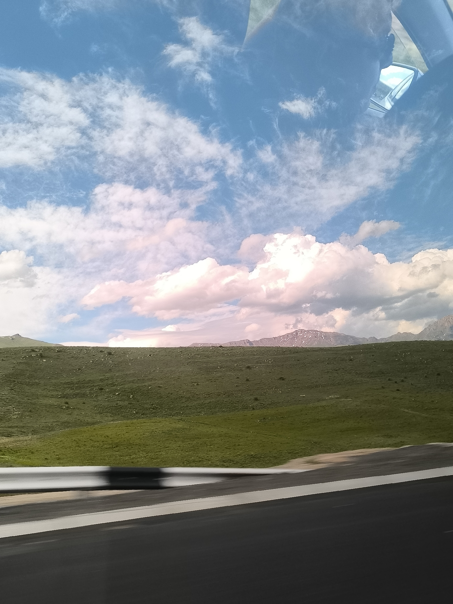 The image presents a scenic landscape captured from the perspective of a moving vehicle in Yelpin, Armenia. The upper two-thirds of the frame are dominated by a vast blue sky adorned with an array of white and pink-hued clouds, ranging from wispy cirrus to large, voluminous cumulus formations. The soft pink tones on some clouds suggest a particular atmospheric condition or possibly the time of day, though the bright overall illumination indicates daylight.
Below the sky, a wide expanse of rolling green hills stretches across the middle ground. These hills are covered in lush, low-lying vegetation, appearing largely treeless and undulating. In the far distance, rugged mountains are visible, some exhibiting lighter patches that suggest snow caps or exposed rock formations, adding to the grandeur of the landscape.
The immediate foreground features elements indicative of travel: a dark asphalt road with a distinct white lane marking on the right, and a white metal guardrail on the left. Both the road and guardrail show signs of motion blur, confirming that the photograph was taken from a vehicle in transit. Reflections are discernible in the upper right corner, likely from a car's windshield or window, including a partial view of a side mirror and a faint outline that could be a person's head or shoulder.
The scene conveys a sense of open-road travel through a beautiful, natural environment, characterized by expansive skies, verdant hills, and distant mountains. The weather appears clear and bright. There is no visible text in the image.