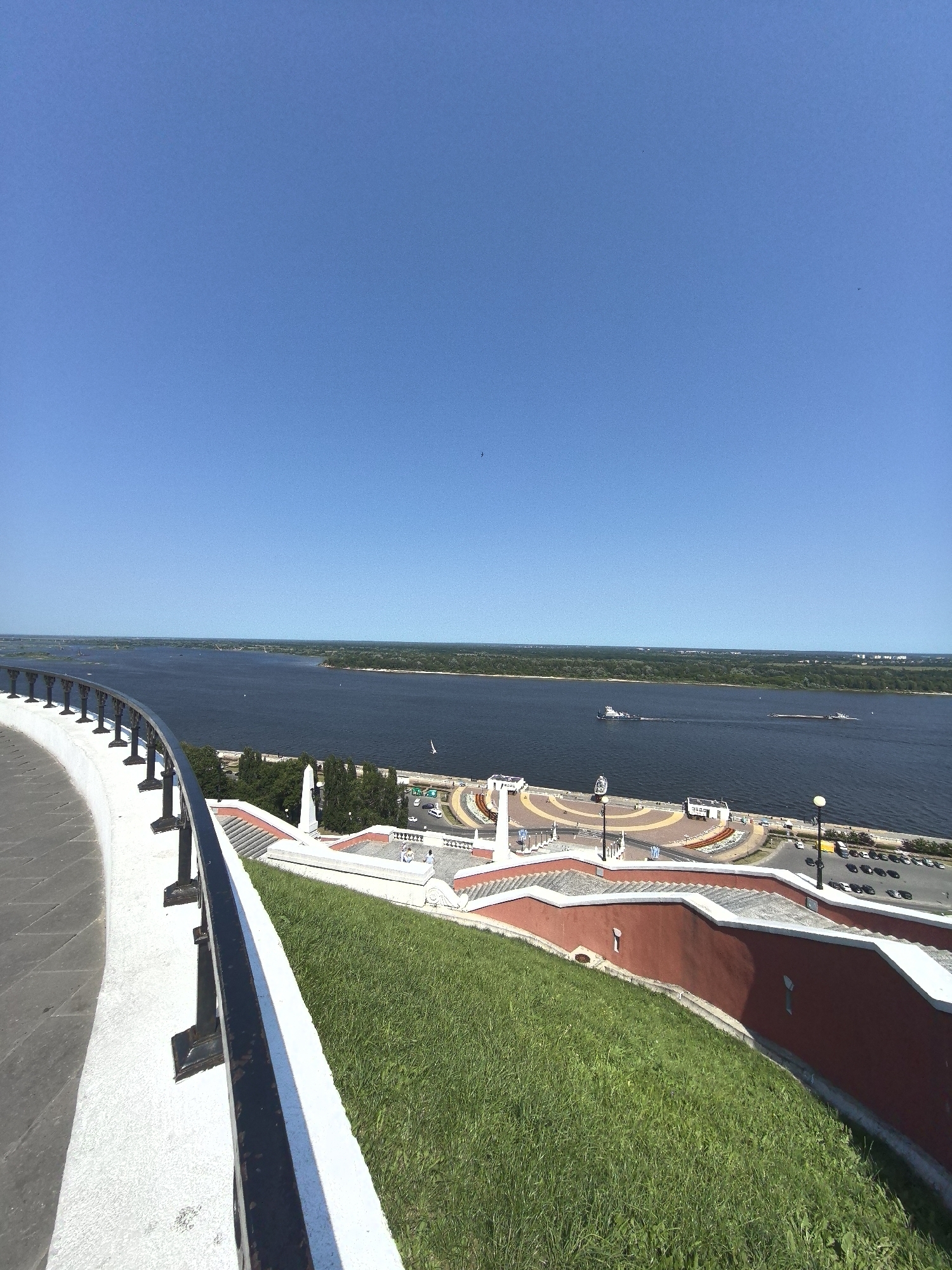 The image captures a scenic view from a high vantage point overlooking the confluence of the Oka and Volga rivers in Nizhny Novgorod, Russia. In the foreground, a white curved wall with a black metal railing lines the edge of a grassy slope. Beyond the railing, a red-colored retaining wall separates the grassy area from a wide, multi-tiered staircase that descends towards the riverfront. The river itself is vast and dark blue, with a distant treelined shore. Several boats, including a tugboat and a barge, are visible on the water. The riverfront features a circular plaza with patterned paving, lampposts, and a few small buildings. The sky above is a clear, vibrant blue with a few faint birds. The perspective suggests this viewpoint is from the Nizhny Novgorod Kremlin or a similar elevated location, offering a panoramic vista of the city's iconic waterways and architectural elements. The scene is bathed in bright daylight, indicating a sunny day.