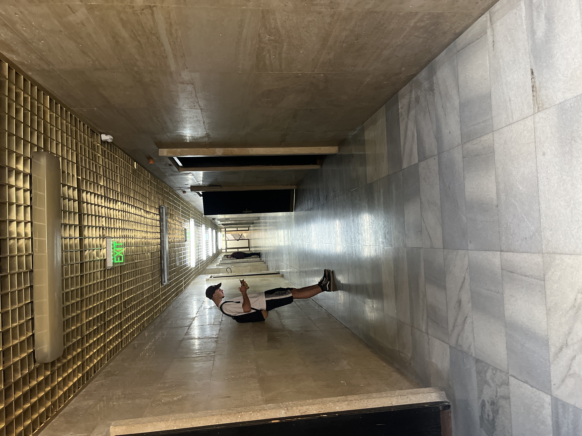 A man lies on his back in a long, narrow indoor hallway. The hallway has walls and a ceiling composed of large, rectangular tiles. One wall is a light grey marble, while the opposite wall features a grid-like pattern of small, metallic gold squares. At the far end of this golden wall is a green "EXIT" sign. The man wears a black baseball cap, a white t-shirt, black shorts with the Adidas logo, and black athletic shoes. He holds a smartphone in his hands, appearing to be looking at it. His feet are propped against the marble wall. In the distance, a second person is visible lying on their back in the same hallway. The scene gives the illusion that the walls are the floor and ceiling, and the man is defying gravity. The hallway appears to be a long, continuous walkway that stretches far into the distance. The architecture and design suggest the interior of a modern building. Given the context, it is likely this photo was taken within the Mall Plovdiv in Plovdiv, Bulgaria.