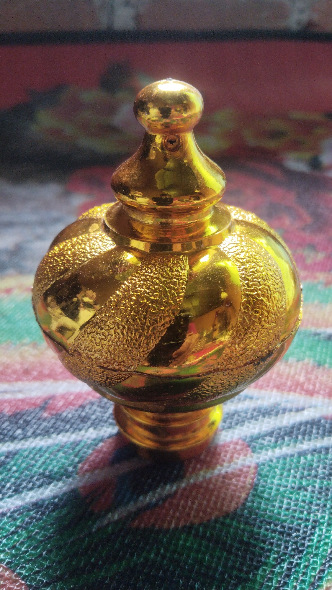 A gold-colored, ornate perfume bottle is the subject of the image. The bottle has a spherical body with a textured, patterned surface, and a decorative cap on top. It rests on a multi-colored, patterned surface that appears to be fabric, possibly a tablecloth or mat. The background is blurred and shows a dark, reddish-brown color with hints of lighter colors. The lighting creates strong reflections on the shiny surface of the bottle. The image is taken from a slightly low angle, looking up at the bottle. This object is likely a decorative item found within a home or shop.