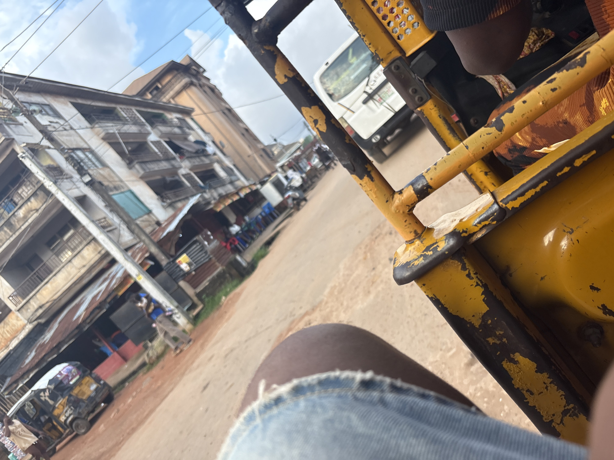 The image depicts a street scene in Onitsha, Nigeria, viewed from the perspective of someone inside a yellow, three-wheeled vehicle, likely a tuk-tuk or auto-rickshaw. In the foreground, parts of the vehicle's yellow frame are visible, with peeling paint and some dark metal underneath. A person's leg, wearing denim shorts, is also in the foreground on the left.
The middle ground shows a busy street with a variety of buildings. On the left, a yellow and black tuk-tuk is parked. The buildings behind it appear to be a mix of weathered structures with corrugated metal roofs and multi-story buildings with balconies. A white truck is visible further down the street, and other vehicles and people are scattered throughout the scene, suggesting a commercial or residential area. The ground is a reddish-brown, characteristic of dirt or unpaved roads in the region.
The background features a large, multi-story building with a weathered facade and balconies. Power lines crisscross the sky, which is partly cloudy. The overall impression is of a vibrant, bustling urban environment typical of Nigerian cities. The perspective suggests the viewer is either driving or a passenger in the foreground vehicle, looking out at the surroundings.