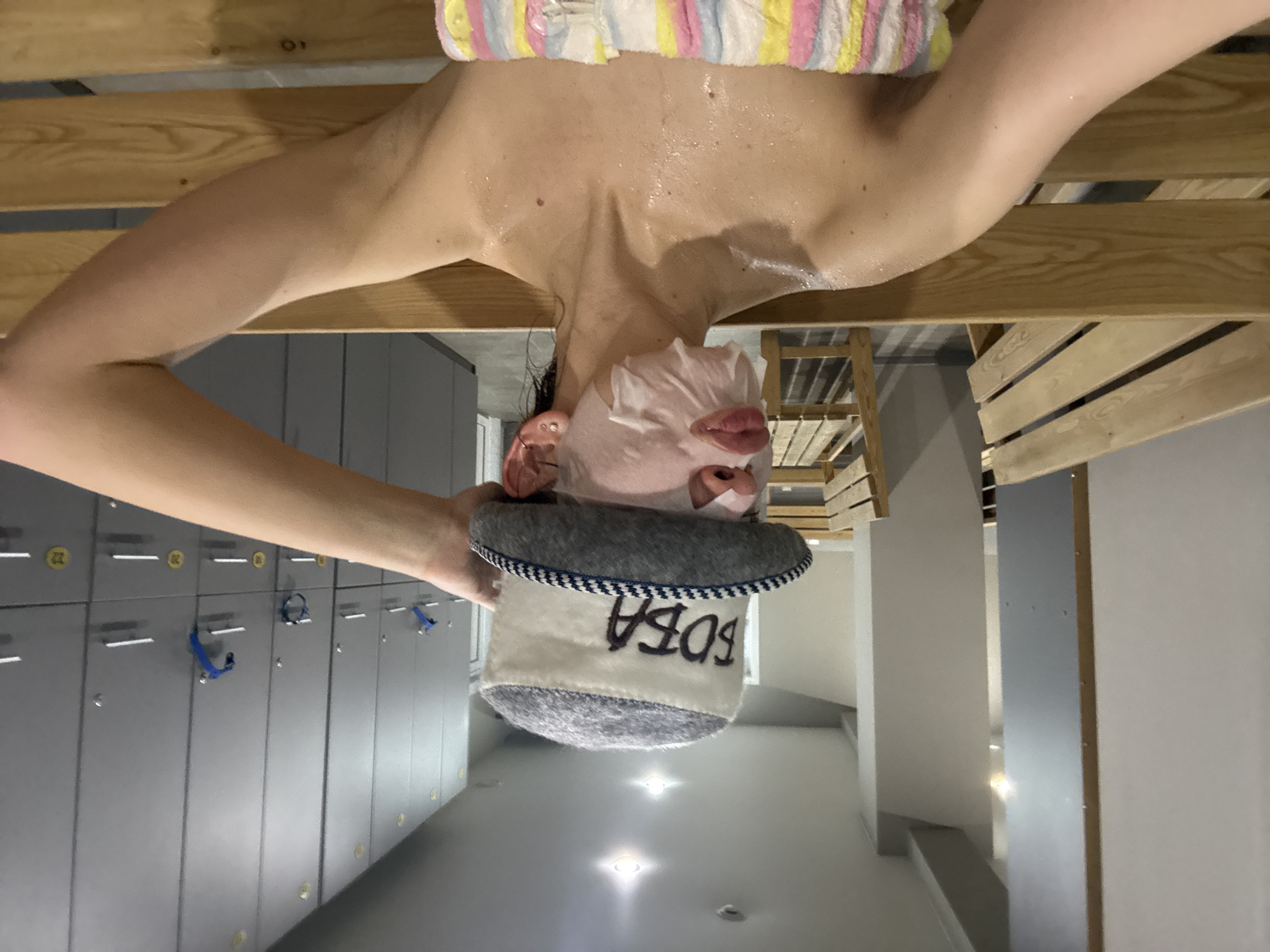 The image shows a woman wearing a face mask and a sauna hat with the word "БОБA" (BOBA) embroidered on it. She is lying on a wooden bench in a sauna or a similar facility. The background includes lockers, lights, and part of the sauna's wooden structure. The location could possibly be a sauna in Ust-Ilimsk, Russia.