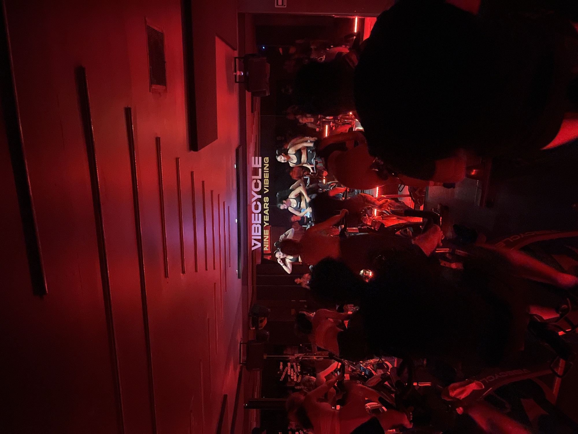 The image captures a dimly lit indoor cycling studio at Vibecycle in San Bernardino Tlaxcalancingo, Mexico. The walls are a deep red, adorned with vertical design elements. A reflective surface or mirror runs along one wall, showing the class in session. Individuals are on stationary bikes, presumably participating in a cycling class. The lighting is predominantly red, creating a low-light environment. The studio appears modern and well-equipped. A sign reads "Vibecycle: Nine Years Vibing."