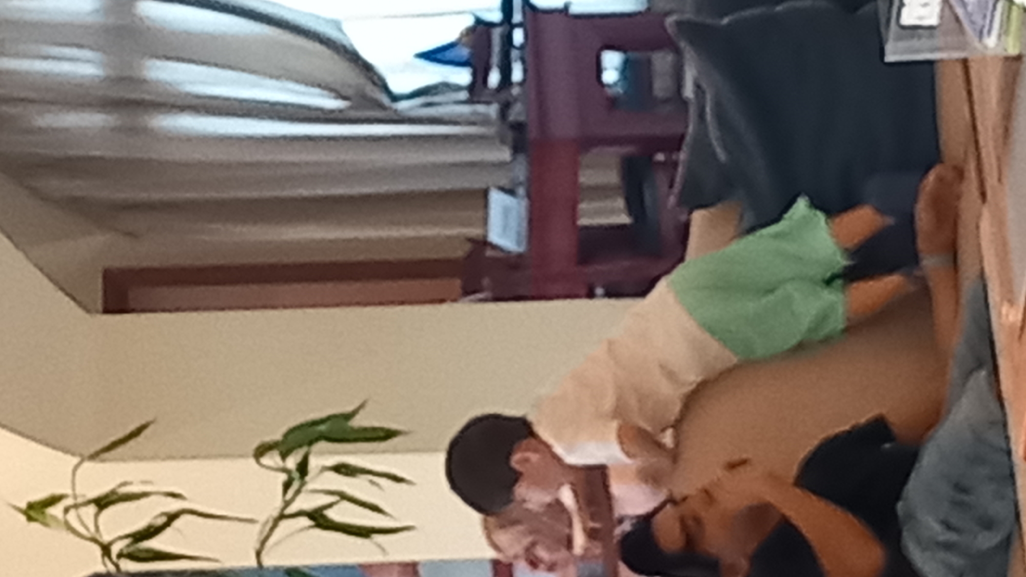 The image shows two people on a surface that appears to be a bed or sofa. The person on the left is a child wearing a white shirt and light green shorts, lying on their side with their legs bent. They have dark hair and are looking towards the other person. The second person, lying on their back, appears to be an adult wearing a dark shirt. Their face is not fully visible, but they seem to be interacting with the child. In the background, there are blurred objects, including what looks like a red rack or shelf and some curtains. The lighting suggests it could be daytime. The overall scene appears to be indoors, possibly a home or hotel room, given the furniture and indoor plants visible in the foreground. There is no visible text in the image.