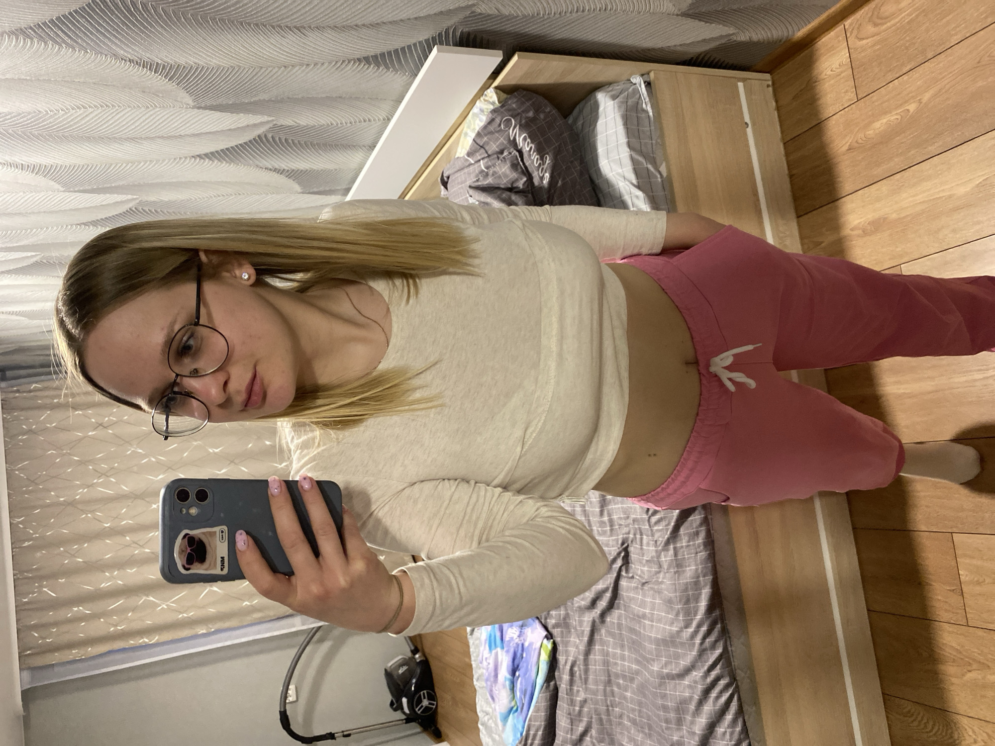 A young woman is taking a selfie in what appears to be her bedroom. She is wearing glasses, a light-colored long-sleeved crop top, and pink sweatpants. She is holding a smartphone with a sticker on the back.
The bedroom has light-colored wooden flooring and a bed with gray and white bedding. There is also a headboard and some kind of cleaning appliance in the room. The wall has a textured wallpaper.
There is no information available from the provided image to indicate that this image was taken in Izhevsk, Russia. The location cannot be determined.