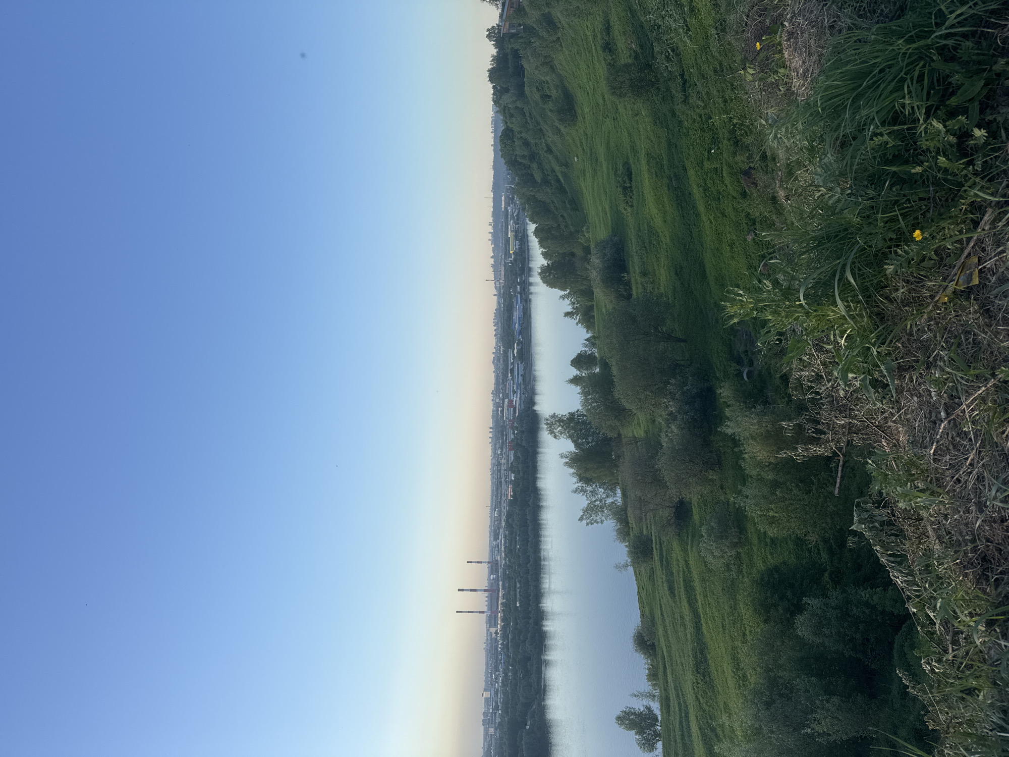 The image shows a panoramic view of the city of Nizhny Novgorod, Russia, specifically a river landscape under a clear, blue sky. A wide river, likely the Volga or Oka, reflects the sky. Along the riverbank, there's a green, grassy slope interspersed with trees and vegetation. In the distance, on the opposite bank, a cityscape is visible, including industrial structures like smokestacks, and a densely populated urban area. The lighting suggests it's either early morning or late afternoon.