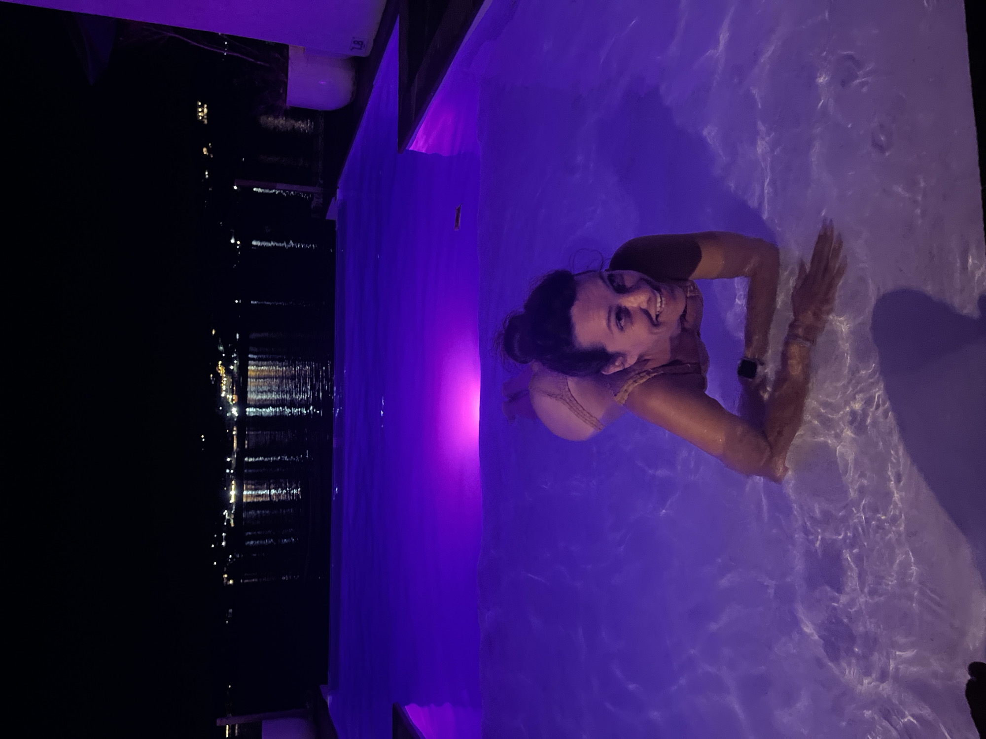 A woman in a tan bikini is submerged in a dimly lit swimming pool at night. The pool is illuminated with purple lights, creating a vibrant glow that reflects off the water. The woman is on her stomach, her head turned towards the camera with a wide smile. She is wearing a watch on her left wrist. In the background, across a body of water, is a silhouette of a city with numerous lights, suggesting an urban coastline. The architecture in the background appears to be a mix of buildings and possibly some vegetation. The location is identified as Nusa Penida, Indonesia.