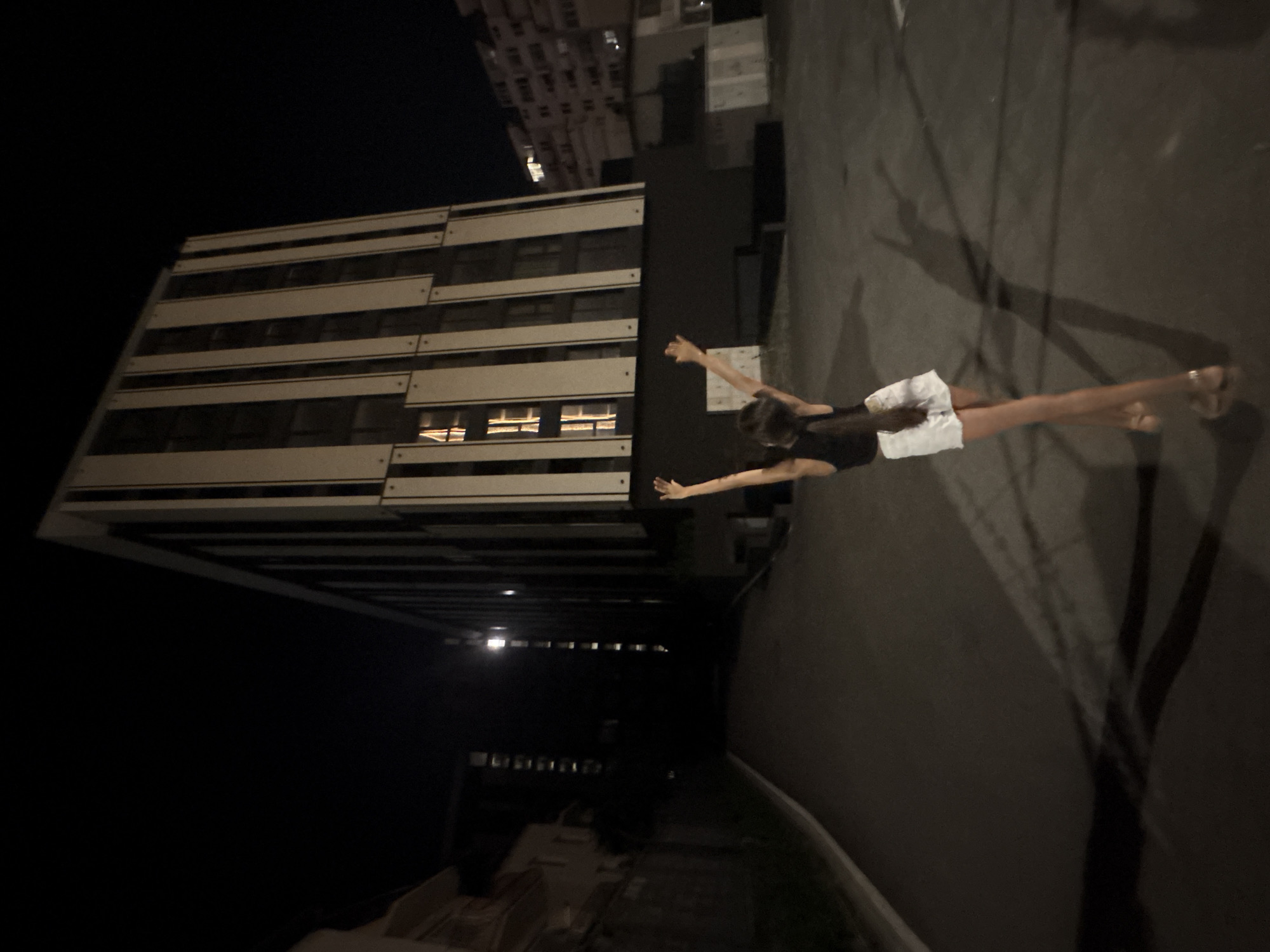 A young person with their back to the camera stands in a dimly lit urban area at night, with their arms raised in a celebratory gesture. They are wearing a dark tank top and white shorts. The surrounding environment features a modern building with vertical white panels and dark windows, a paved pathway or road, and other indistinct buildings in the background. The scene is illuminated by artificial lights, casting long shadows.