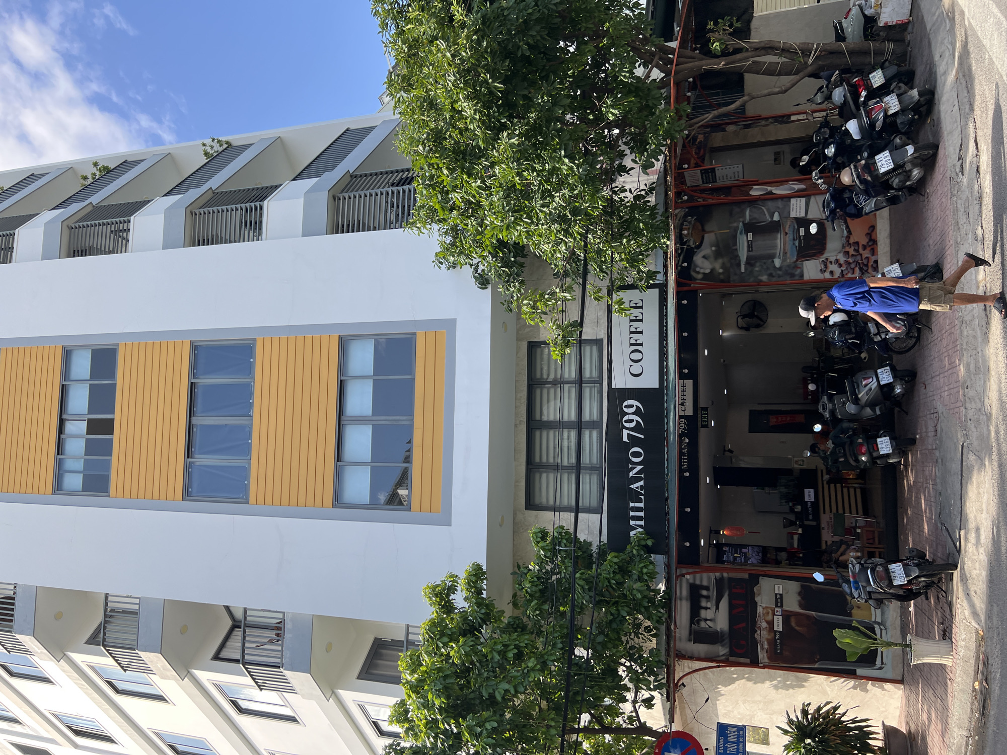 In Nha Trang, Vietnam, a tall, modern building with a light grey facade and orange accents rises against a clear blue sky. The building features multiple floors with balconies and windows. At the base of the building, a cafe named "MILANO 799 COFFEE" is visible. The cafe has a dark awning and large windows displaying artwork of coffee drinks and coffee beans. Several motorbikes are parked in front of the cafe. A man wearing a blue t-shirt and khaki shorts is walking past the cafe on the sidewalk. The street in front of the cafe is paved with pinkish bricks. A traffic sign, a plant in a white pot, and a large green tree with lush foliage are also present on the sidewalk.
