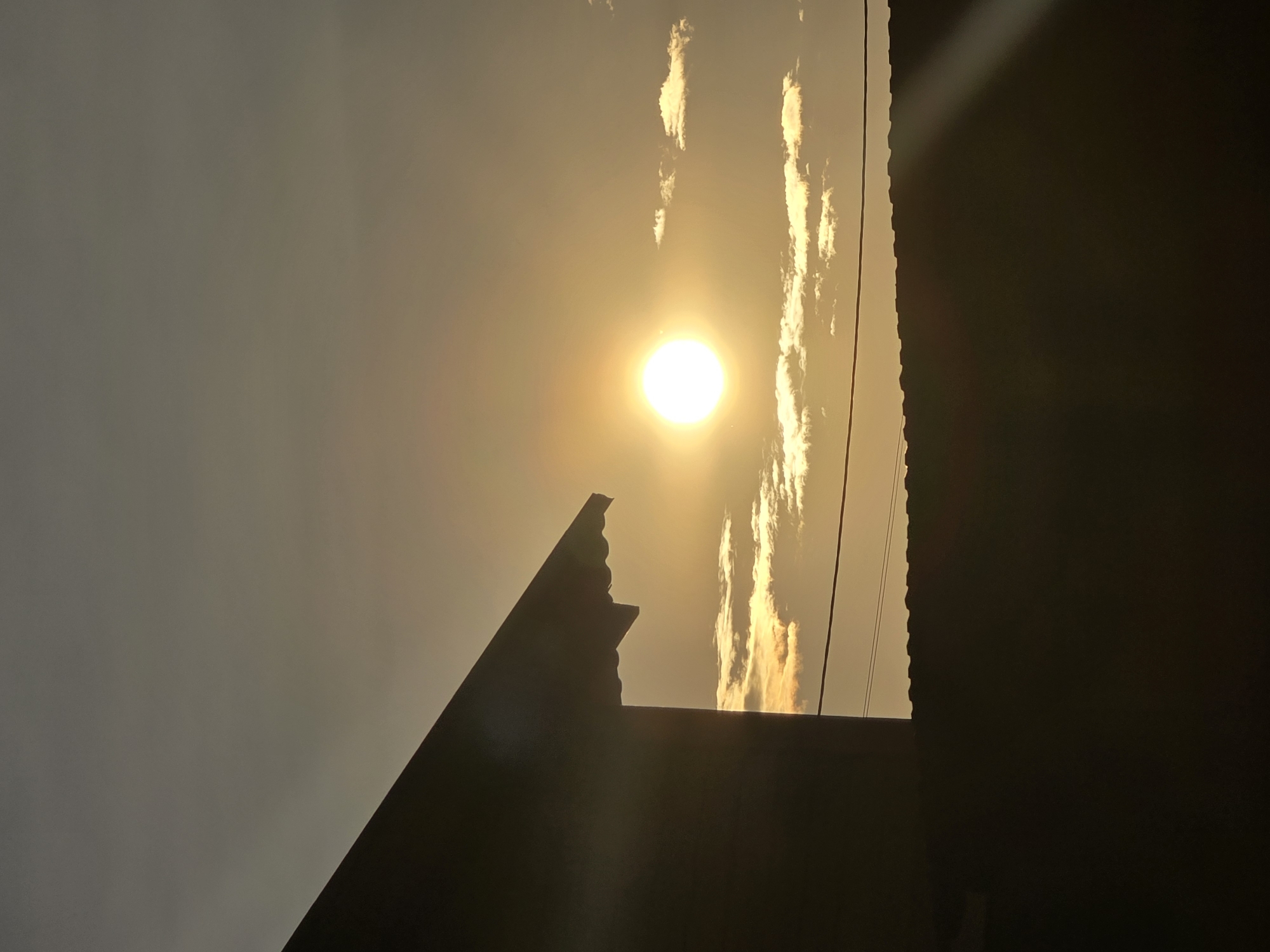 The image captures a silhouette of a building against a sunset sky. The sun, a bright, glowing orb, is positioned towards the center of the frame, casting a warm, golden light. Wisps of clouds are scattered across the sky, illuminated by the setting sun. In the foreground, the sharp, angular outline of a building's roof and wall is visible, rendered in dark shadow. The overall scene suggests a peaceful evening setting. The location is identified as Ермолаево, Russia. There are no people or animals present in the image. The image focuses on the architectural elements and the natural phenomenon of sunset.