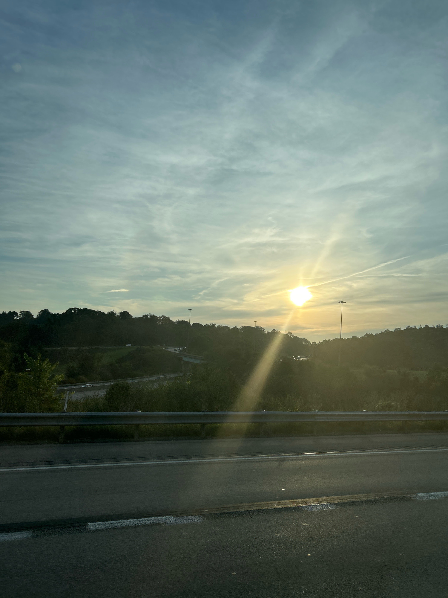 The image depicts a scenic view of a highway, bordered by a metal guardrail, under a partially clouded sky. The sun is prominently visible in the upper right quadrant, casting a warm glow and creating visible sunbeams that stretch across the frame. The landscape features rolling hills covered in lush green trees, with a winding highway visible in the mid-ground, populated by a few vehicles. A tall light pole stands near the sun, also silhouetted against the bright sky. The road in the foreground appears to be wet, reflecting the soft light of the sun. The overall impression is one of a serene, possibly early morning or late afternoon vista on a highway. The location is identified as Robinson Township, USA.