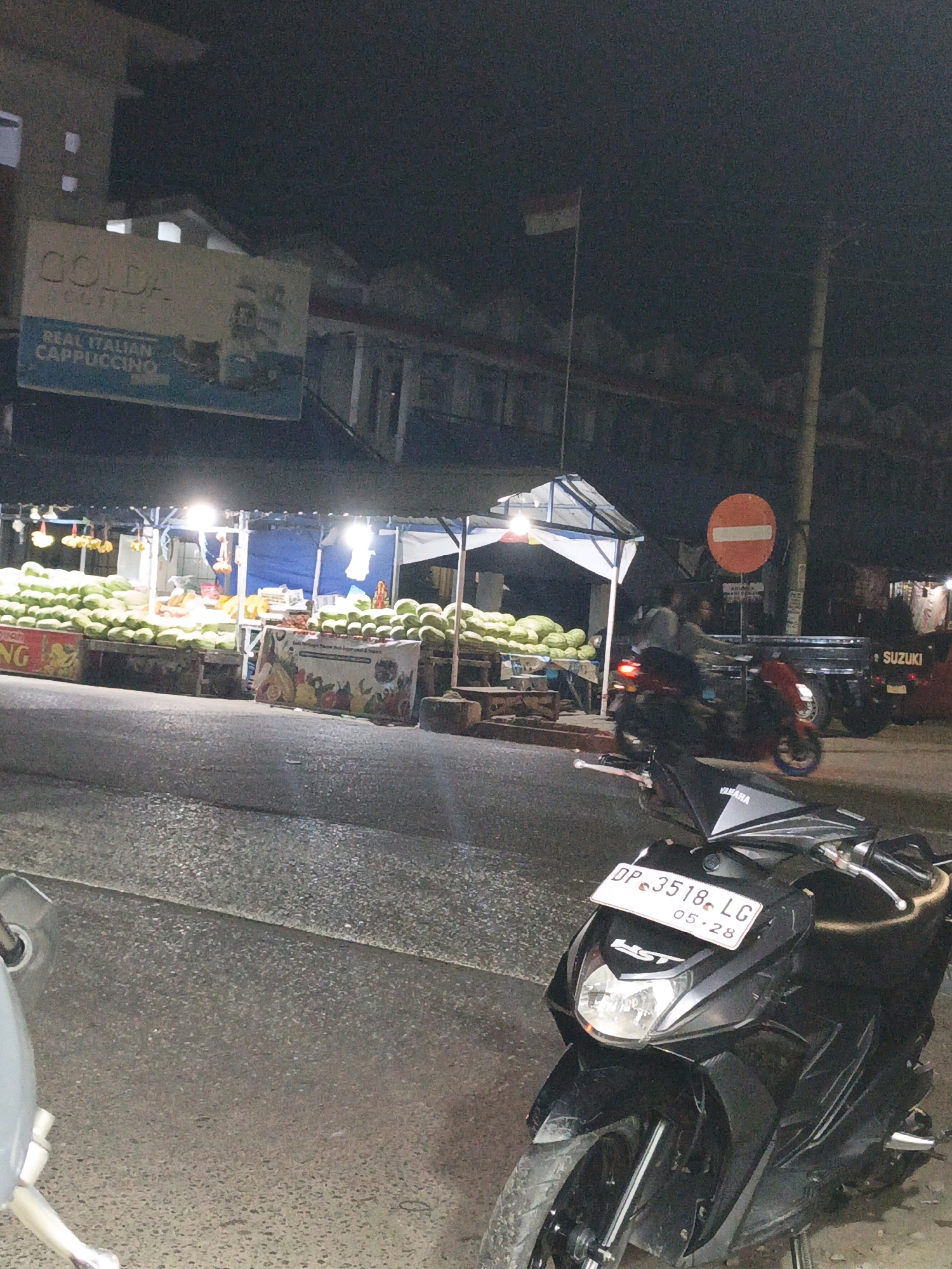 At night, in the city of Pangkajene, Indonesia, a street-side fruit stand is illuminated by artificial lights. Large piles of green fruit, possibly watermelons, are displayed on tables and the ground. In the foreground, a black Yamaha scooter is parked, its license plate reading "DP 3518 LG". Behind the scooter, a red Suzuki pick-up truck is visible, and further down the street, another motorcycle is in motion. A red and white traffic sign with a white bar is mounted on a pole. A flag, likely the Indonesian flag, flies from a pole on a building in the background. A sign for "GOLD. COFFEE REAL ITALIAN CAPPUCCINO" is mounted on the facade of a building. The scene suggests a bustling local market or roadside vendor area.