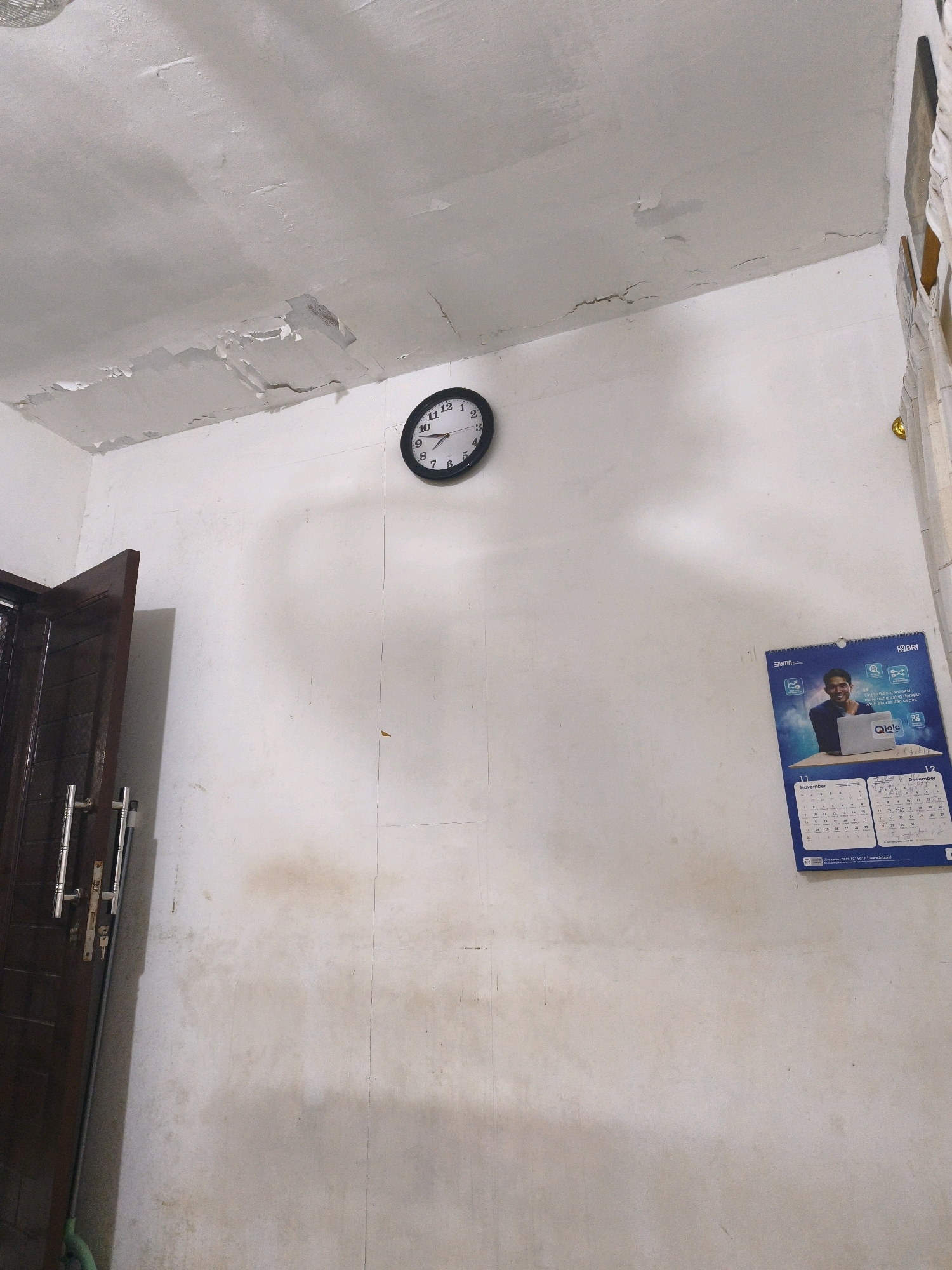 A round analog clock with a black frame is mounted on a white wall. The clock's hands indicate it is approximately 9:47. To the right of the clock, a wall calendar hangs, featuring a picture of a man with a laptop. The calendar shows the months of November and December. To the left of the clock, a dark brown door with silver handles is partially visible. The wall and ceiling show signs of water damage, with peeling paint and stains. A mop leans against the wall near the door. The scene appears to be an interior room, likely in a residential or office setting. Without further context or identifiable landmarks, the specific location within Samarinda, Indonesia, cannot be determined.