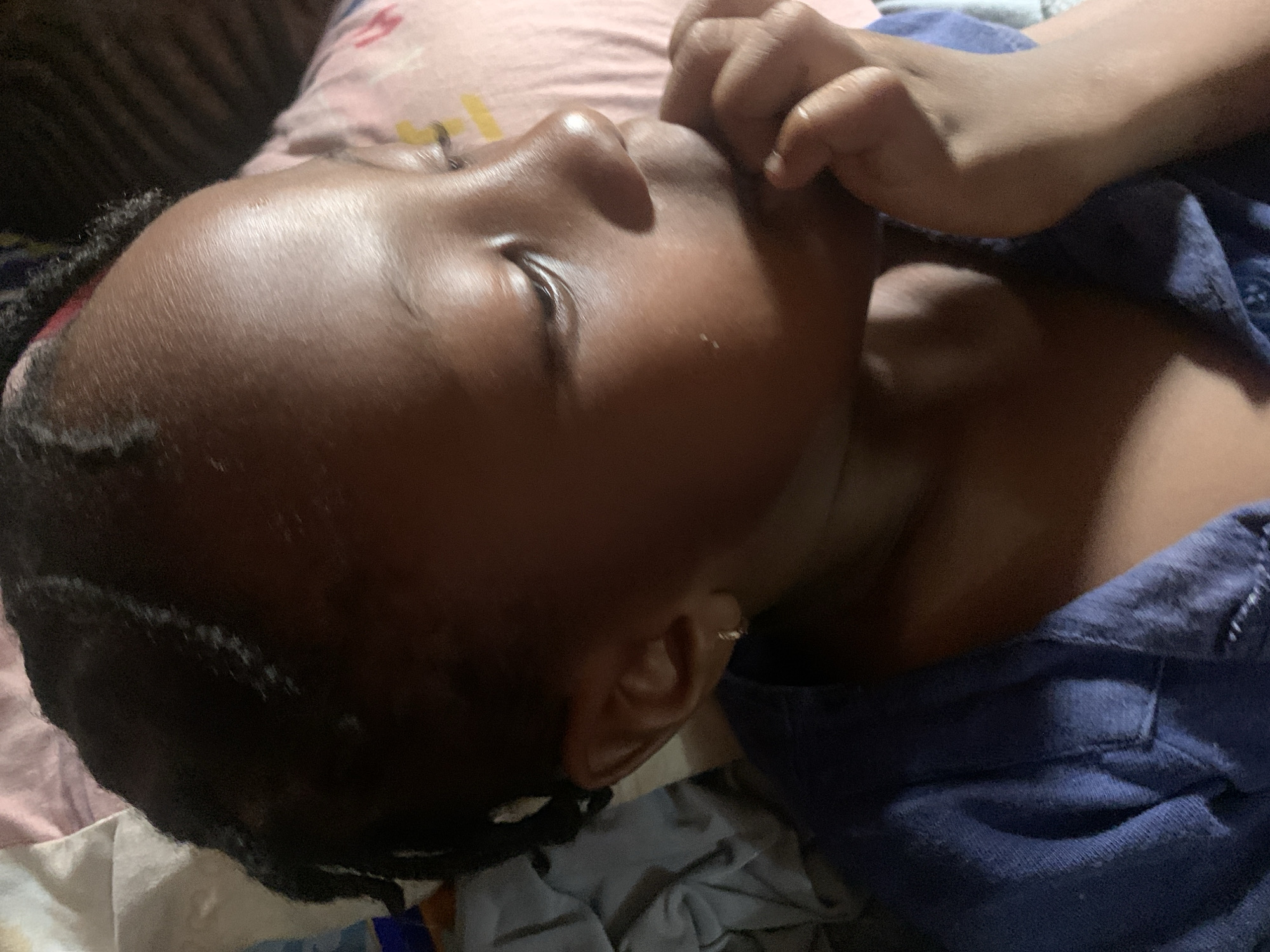 The image shows a close-up, overhead view of a young Black child lying down, resting their head on a pink and white patterned pillow. The child is wearing a dark blue garment. Their face is turned to the right, with their hand near their mouth. The background consists of bedding and a blue cloth. The scene is indoors, likely in a residential setting within Sabon Tasha, Nigeria. The lighting is dim, suggesting an indoor environment at night or in low light conditions. There are no visible external elements that could indicate a specific outdoor location in Sabon Tasha, Nigeria.