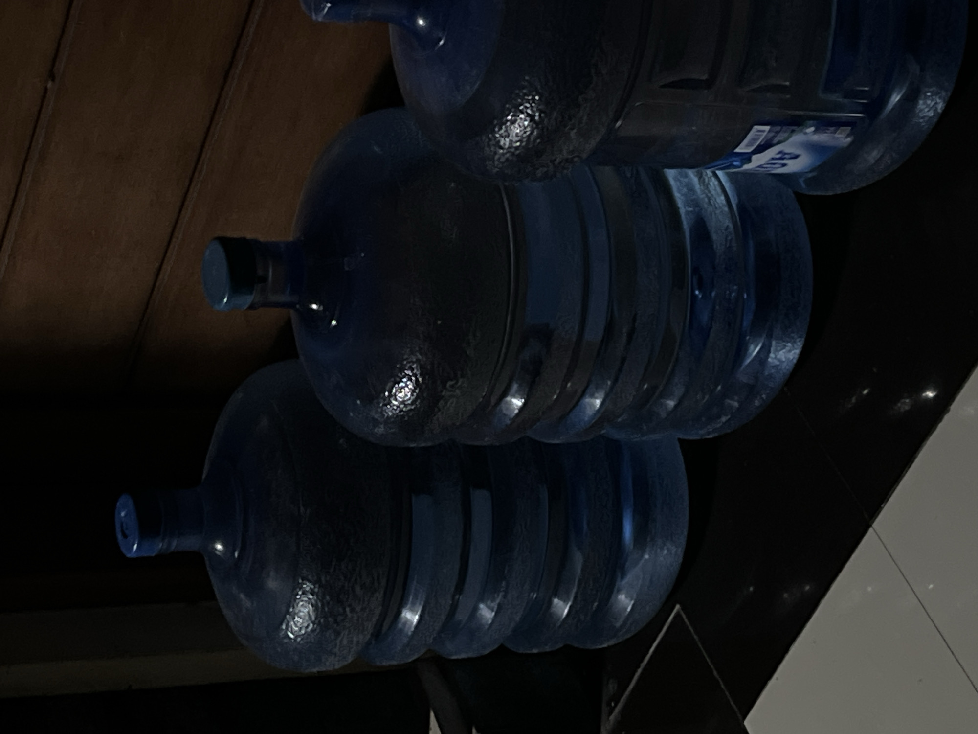The image shows three large, blue water bottles stacked one above the other. The bottles are made of transparent plastic and have textured surfaces. The background on the left appears to be wooden, while the background on the right transitions into black and white tiled flooring. The lighting suggests an indoor environment. There is no clear evidence of a specific location in Canggu, Indonesia. There are no people or actions visible.