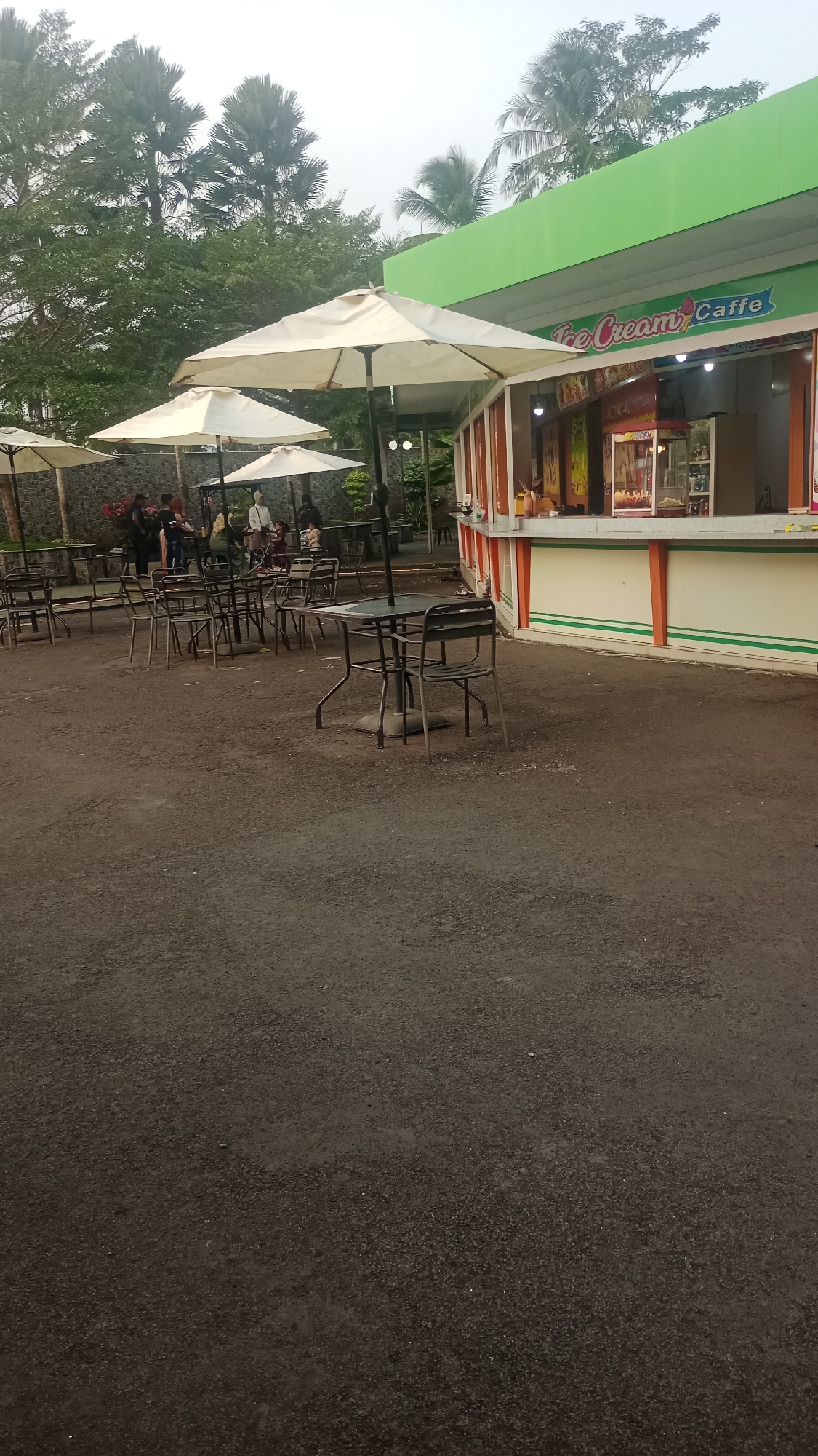 In Pakemitan, Indonesia, an outdoor seating area is depicted in front of an "Ice Cream Caffe" establishment. Several white umbrellas provide shade over tables and chairs arranged on a dark asphalt surface. A few patrons are visible seated and standing around some of the tables. Behind the seating area, the "Ice Cream Caffe" building has a green awning and an open service counter displaying various items. Lush green trees and vegetation form the backdrop of the scene, with palm trees and other foliage visible against a slightly hazy sky. The image captures a relaxed, casual atmosphere at a food and beverage vendor.