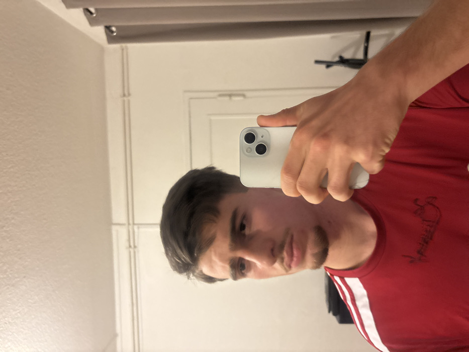 A man in a red shirt with white stripes on the shoulder takes a selfie in a mirror. He holds a white smartphone in his right hand. The background features a white wall with a door. A curtain rod is visible at the top of the frame. The man has dark hair and a trimmed beard. He looks directly into the camera.
