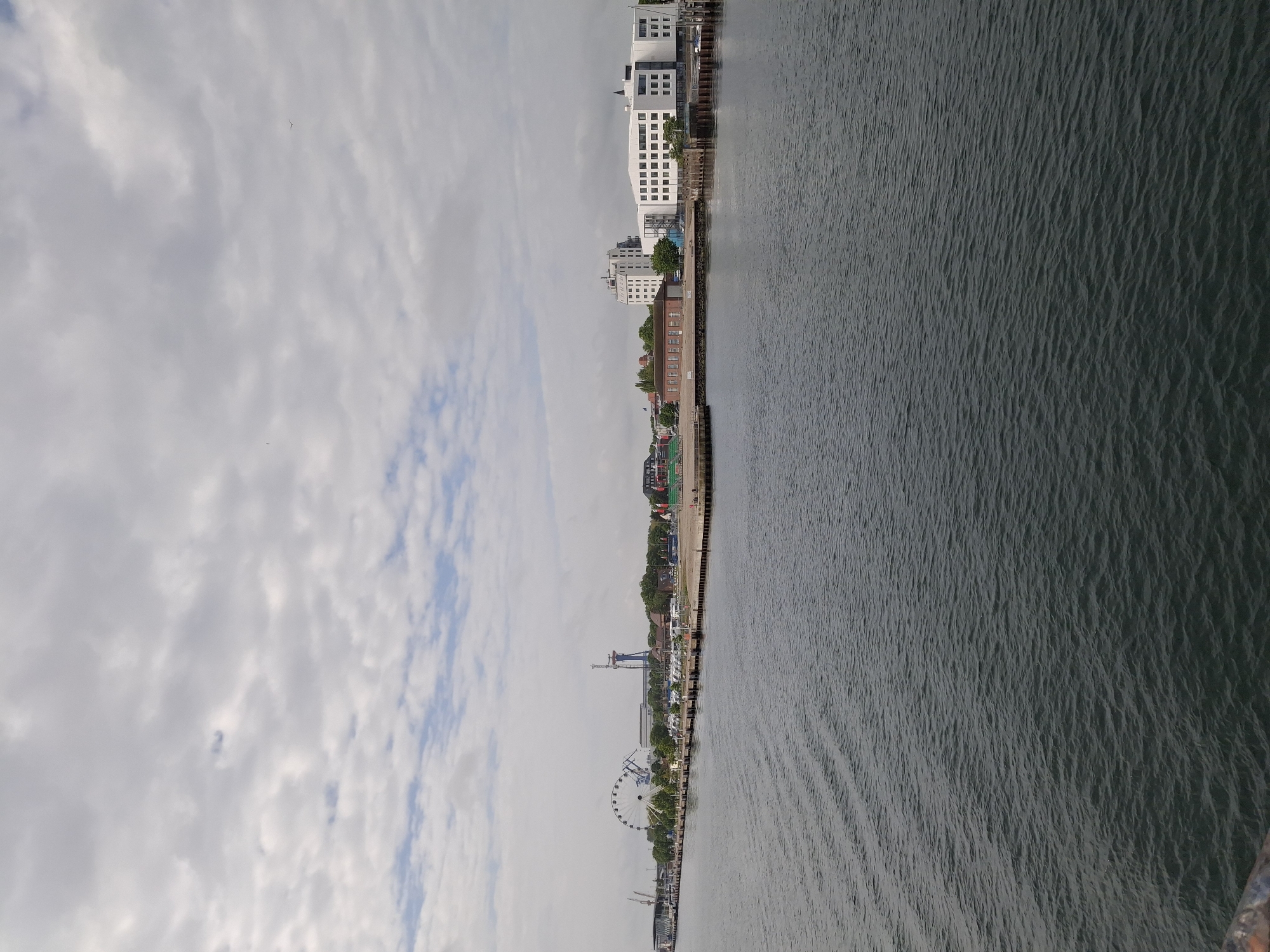 The image shows a waterfront view in Rostock, Germany, featuring a body of water, likely the Warnow River or a harbor area. Along the waterfront are various buildings, including modern white structures and older brick buildings, suggesting a mix of architectural styles. A Ferris wheel is visible, likely part of a recreational or tourist area. The sky is overcast with clouds. The water has visible ripples and wave patterns. The image suggests a typical cityscape along the waterfront in Rostock.